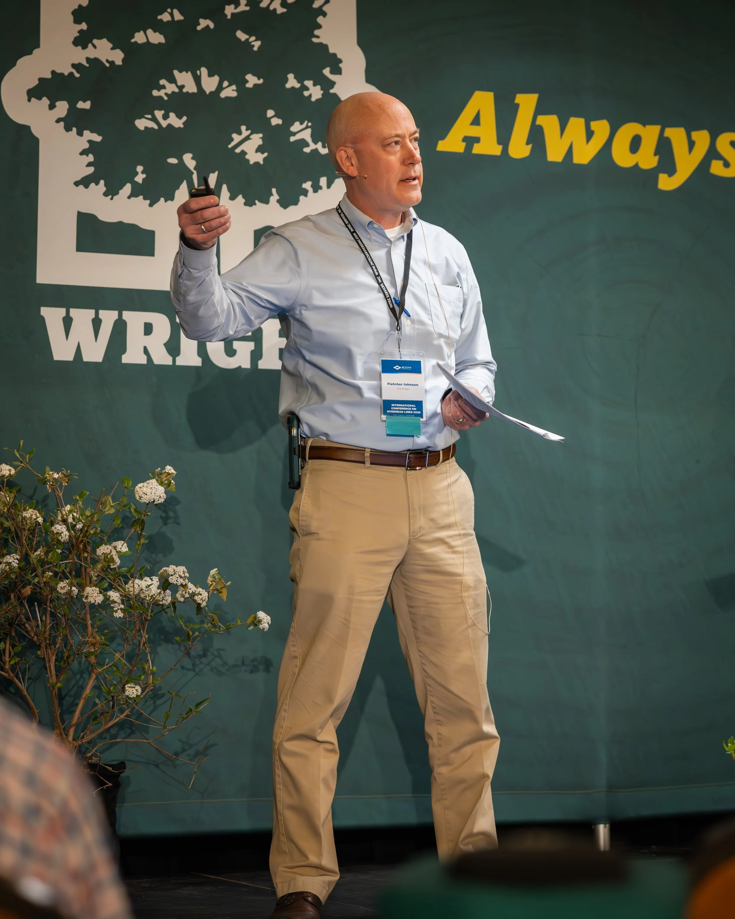 A man with a bald head and light skin, wearing a light blue shirt and beige pants, is standing on a stage giving a presentation. He has a label hanging around his neck that reads 'Fletcher Johnson' and 'International Conference on Overhead Lines 2023