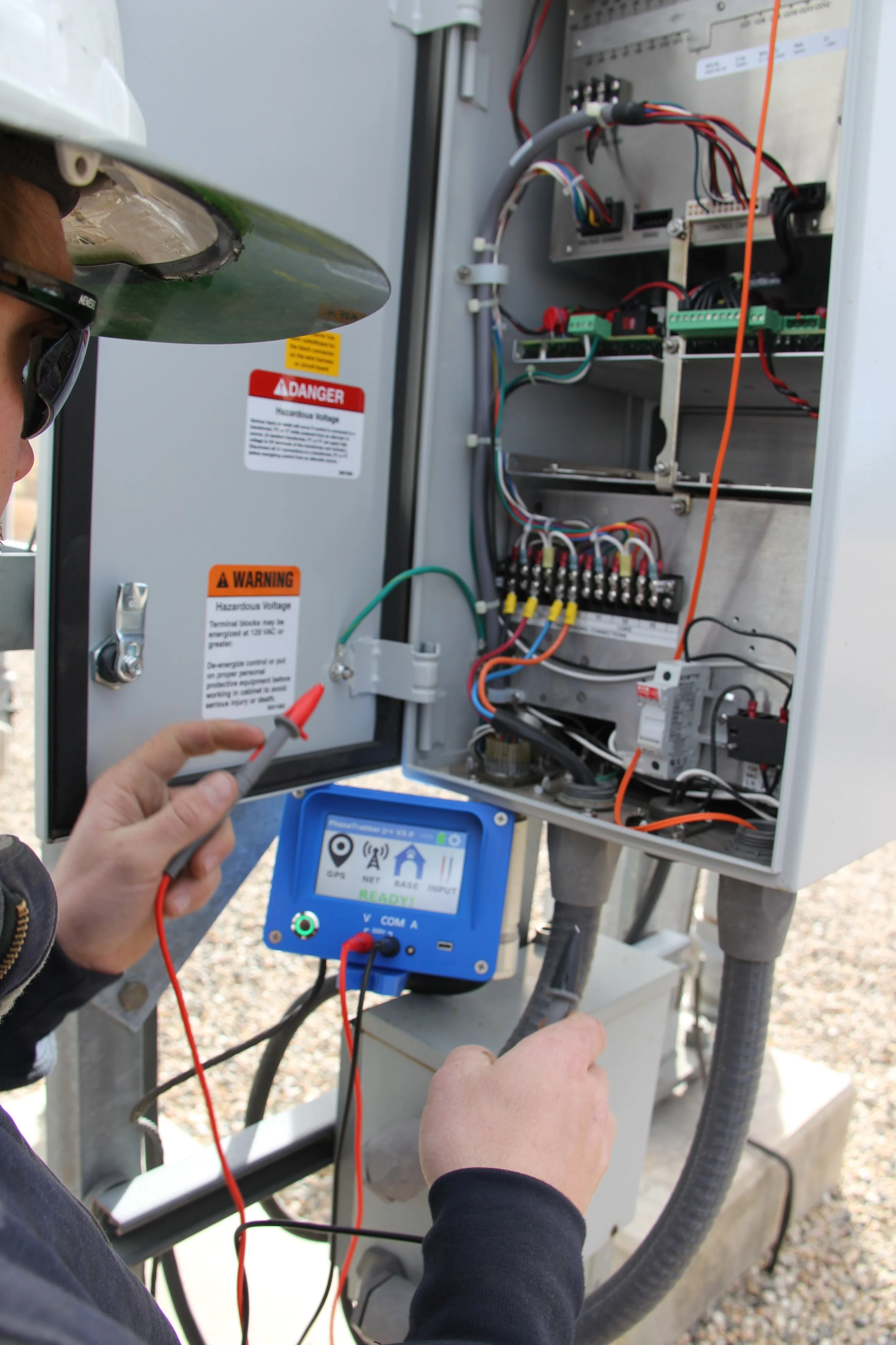 A technician performing maintenance or inspection on an outdoor electrical or telecommunications cabinet, using testing equipment and holding a screwdriver.