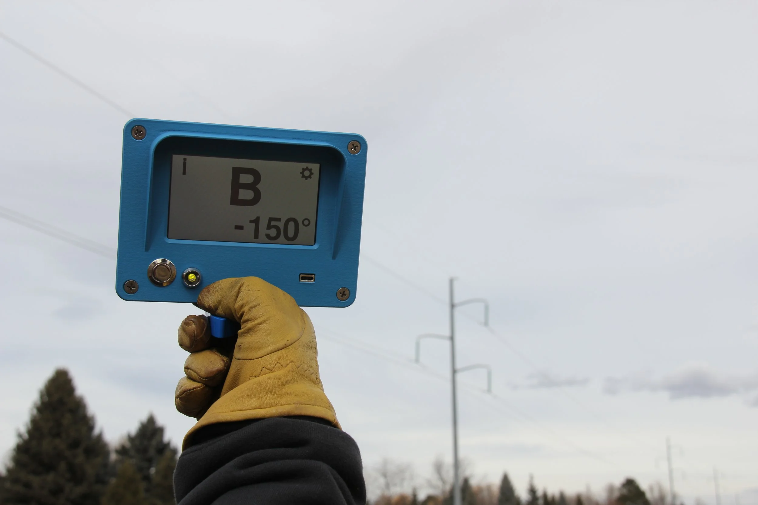 A person wearing a yellow glove holds a digital device displaying altitude and angle measurements against a cloudy sky, with utility poles and trees in the background.
