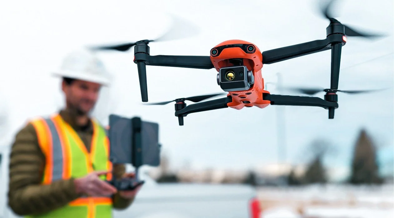 A drone with a camera flying in the air while a man in a construction vest and hard hat operates a remote controller in the background.