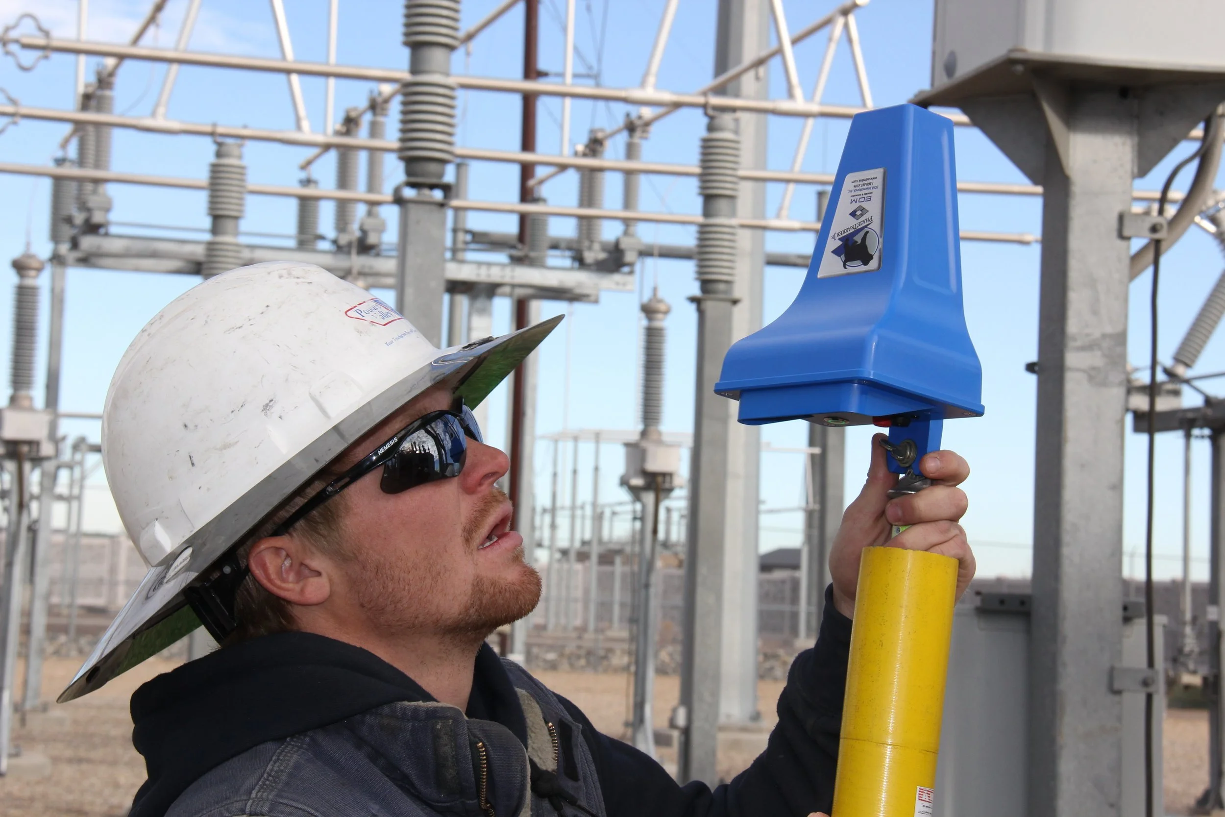 An electrician wearing a white safety helmet and black sunglasses working outdoors at a substation, adjusting a blue electrical device attached to a yellow pole.