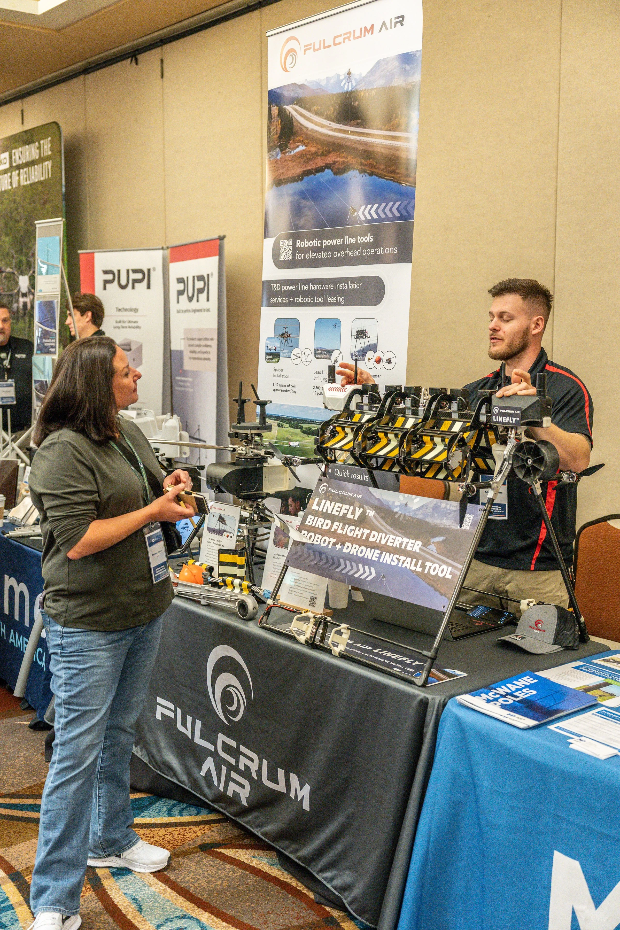 An indoor exhibition booth for Fulcrum Air showcasing drone technology. A woman is standing in front of the booth, engaging with a man behind the table who is explaining drone equipment. The booth displays promotional banners and various drone compon