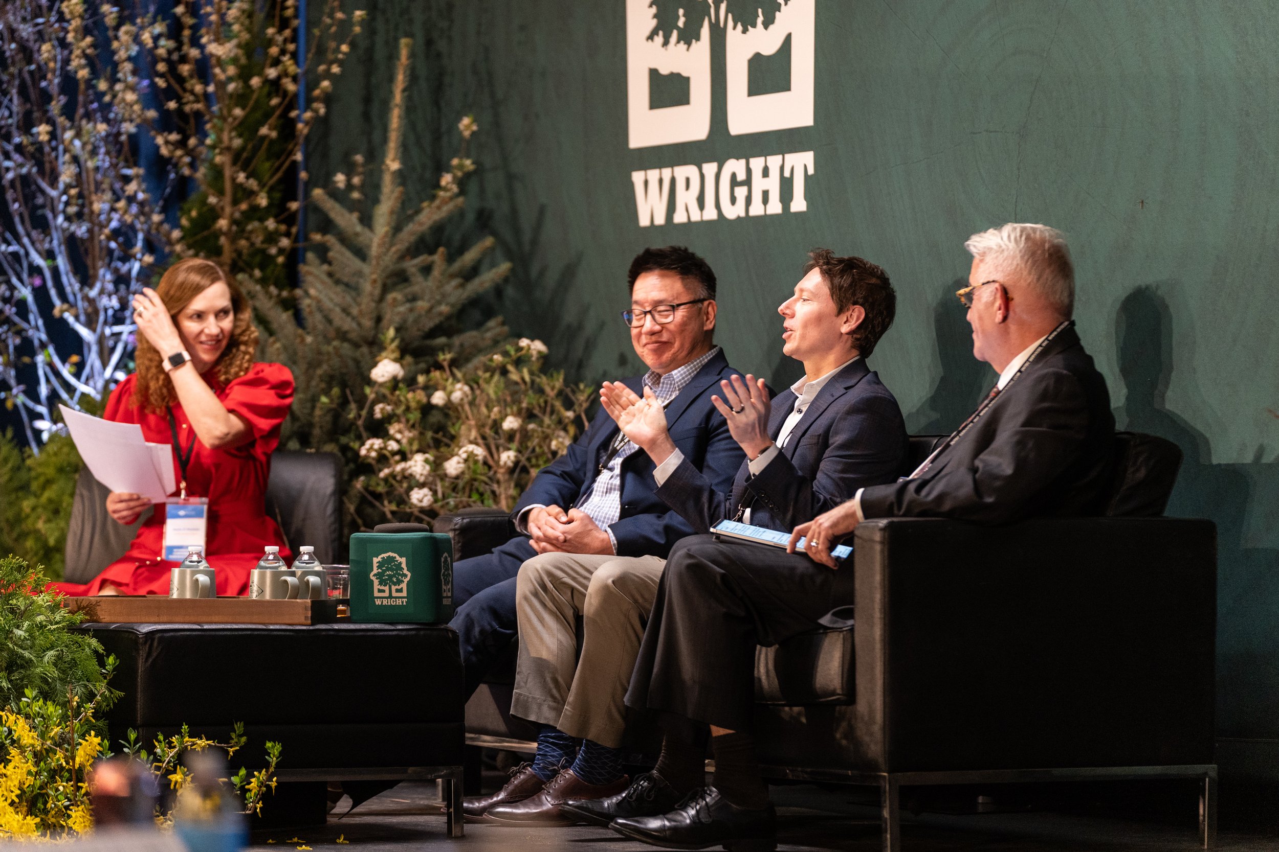 Four people on a panel at a conference, sitting on stage, with a woman in a red dress holding papers, and a green sign with a tree logo that says 'WRIGHT.' There are flowers and decorations around the stage, and a large green background with the word