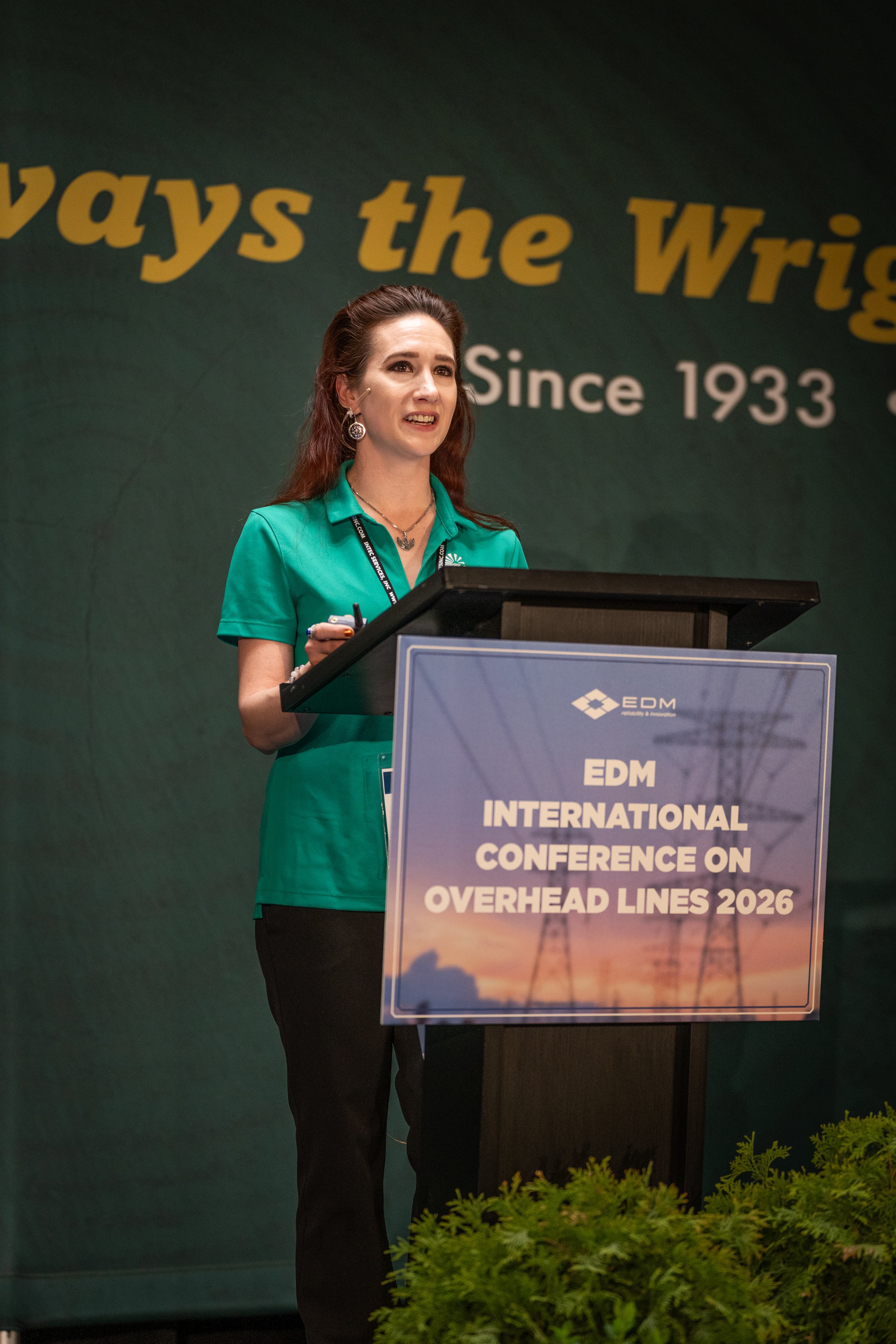 A woman stands at a podium speaking at the EDM International Conference on Overhead Lines 2026, with a green backdrop showing the conference's name and date, wearing a teal top and black pants.