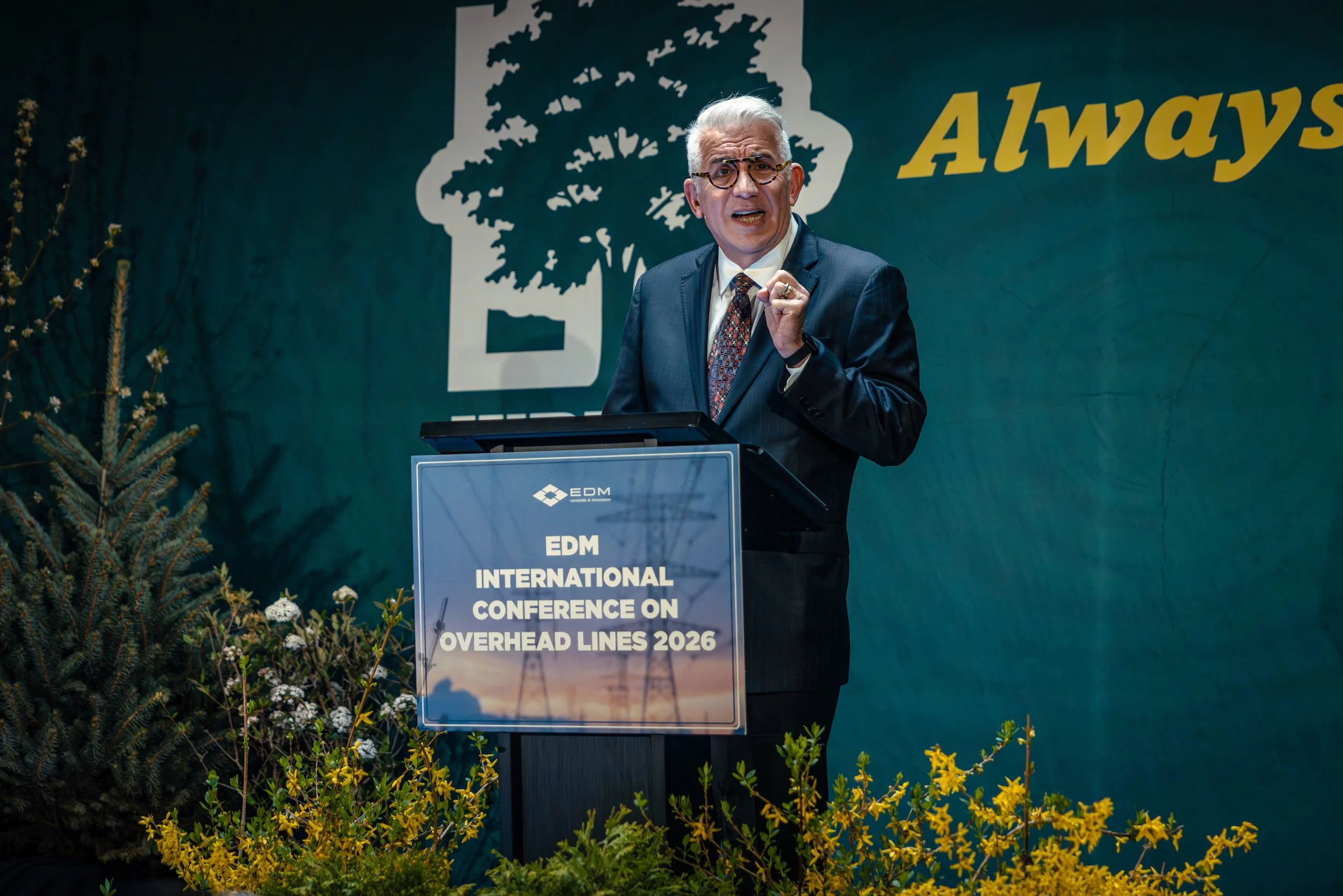 A man in a suit and glasses speaking at a podium during the EDM International Conference on Overhead Lines 2026. Behind him is a large blue backdrop with a white outline of a tree and part of the word 'Always' visible. The podium has a sign with text about the conference, and there are plants and yellow flowers in the foreground.