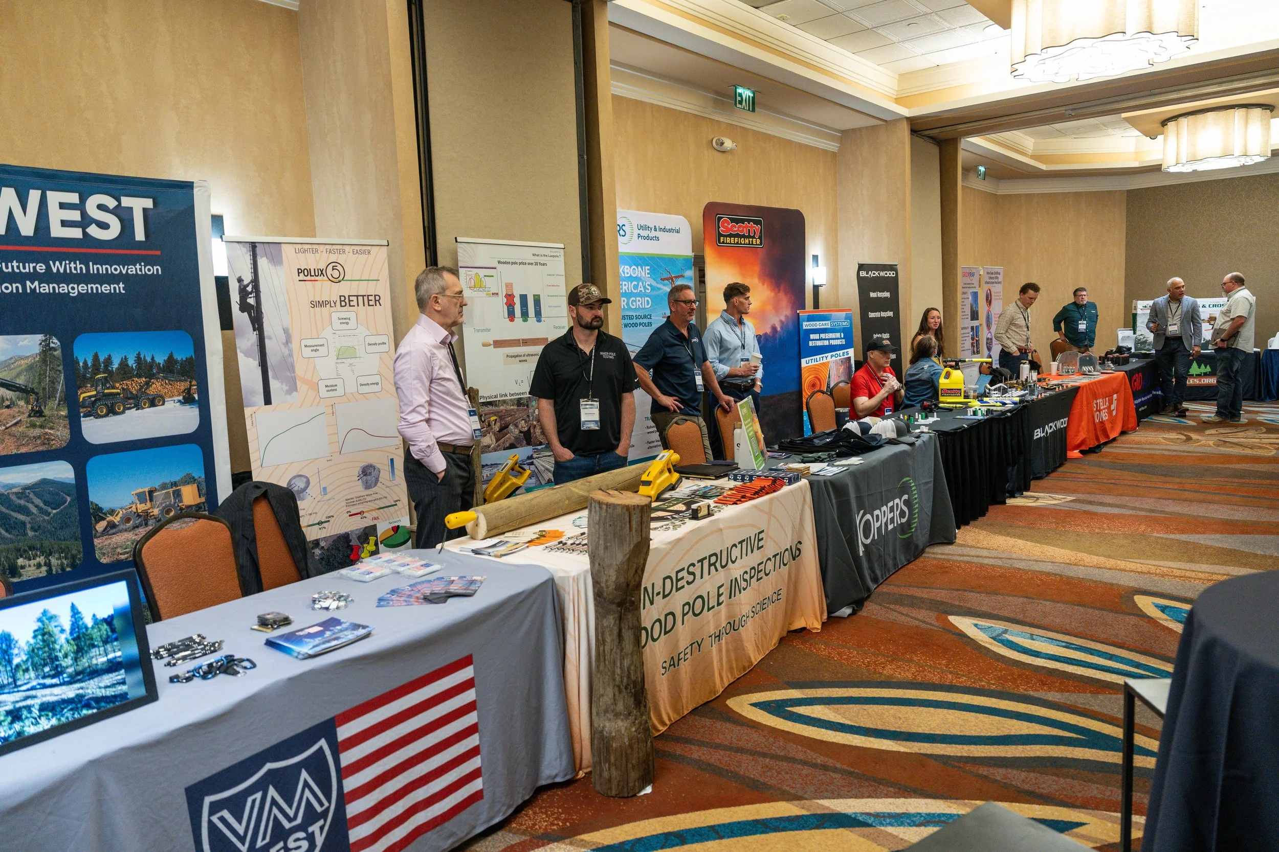 A conference or trade show with multiple booths displaying various products and informational posters. Several people are standing behind the booths, engaging with visitors. The booths have banners and marketing materials related to construction, woo