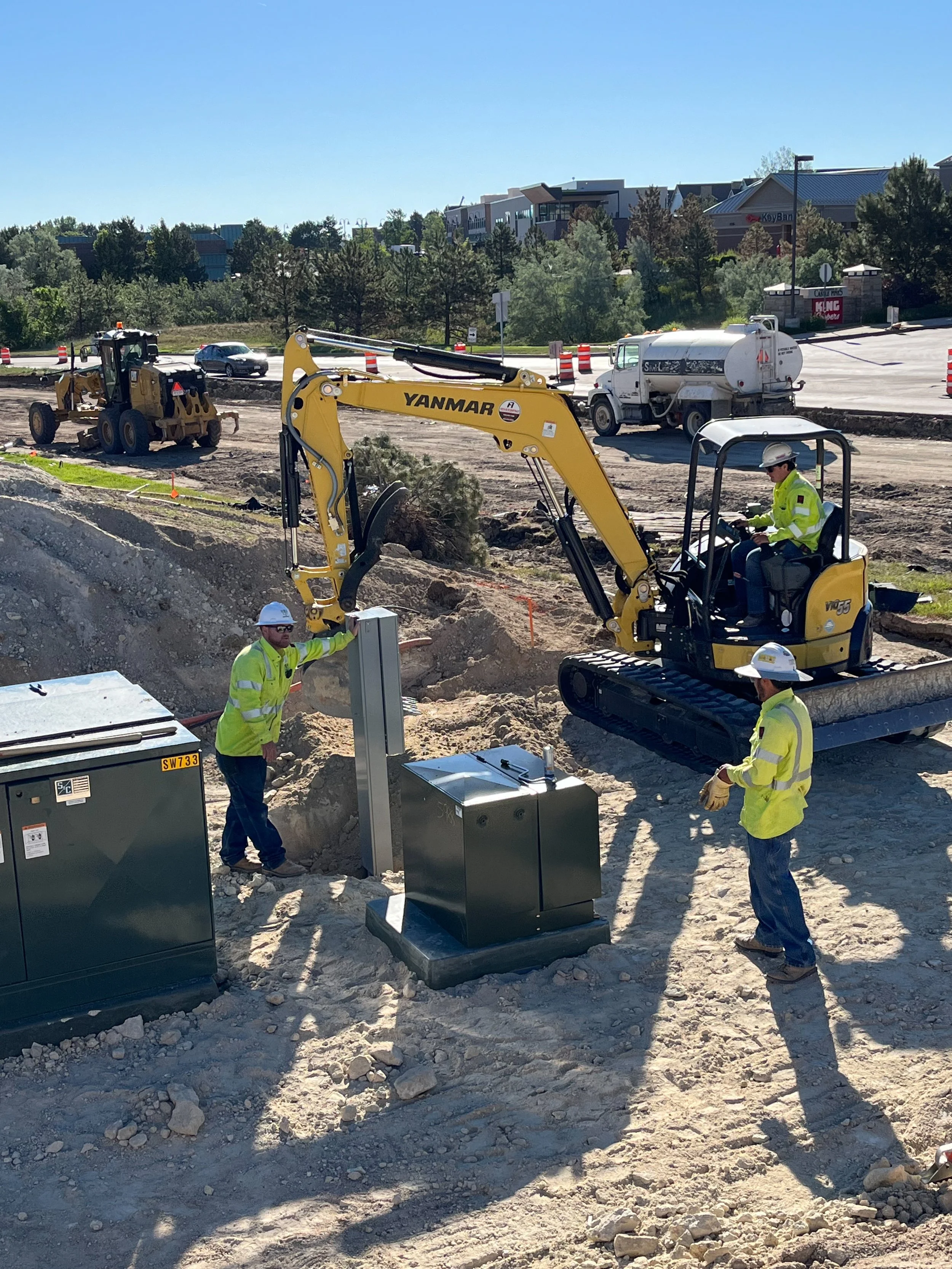 Construction site with workers and machinery installing equipment, with a highway and city buildings in the background.