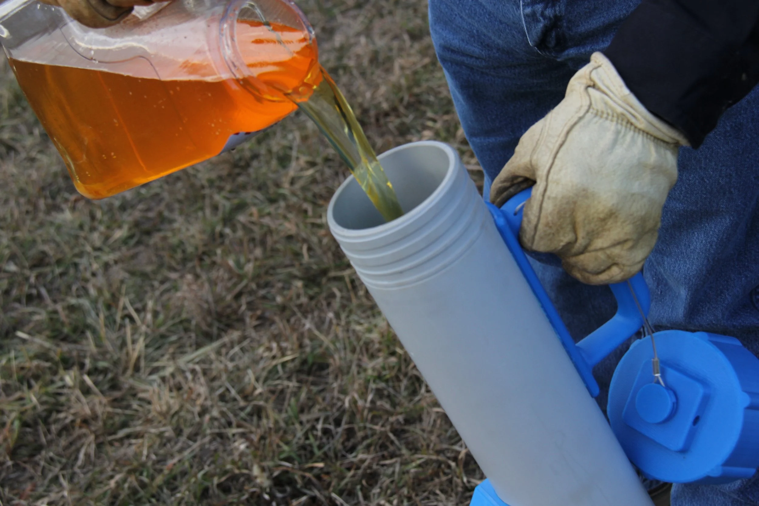 Person wearing work gloves pouring amber-colored liquid from a plastic bottle into a white insulated travel mug, outdoors on grass.
