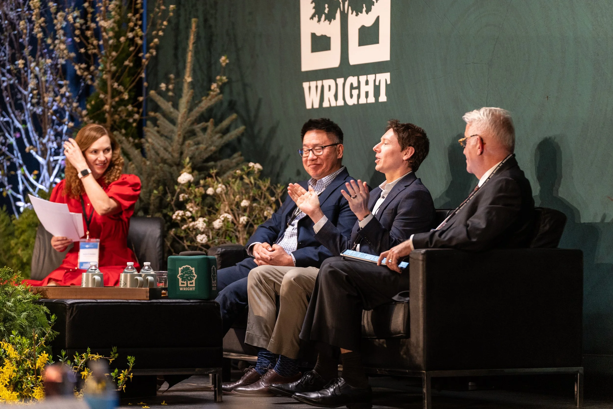 Four people sitting on a stage at a conference or event, with a woman in a red dress standing to the left, holding papers. The background features a large green wall with the word 'WRIGHT' and a logo, and there are decorative trees and plants around the stage.