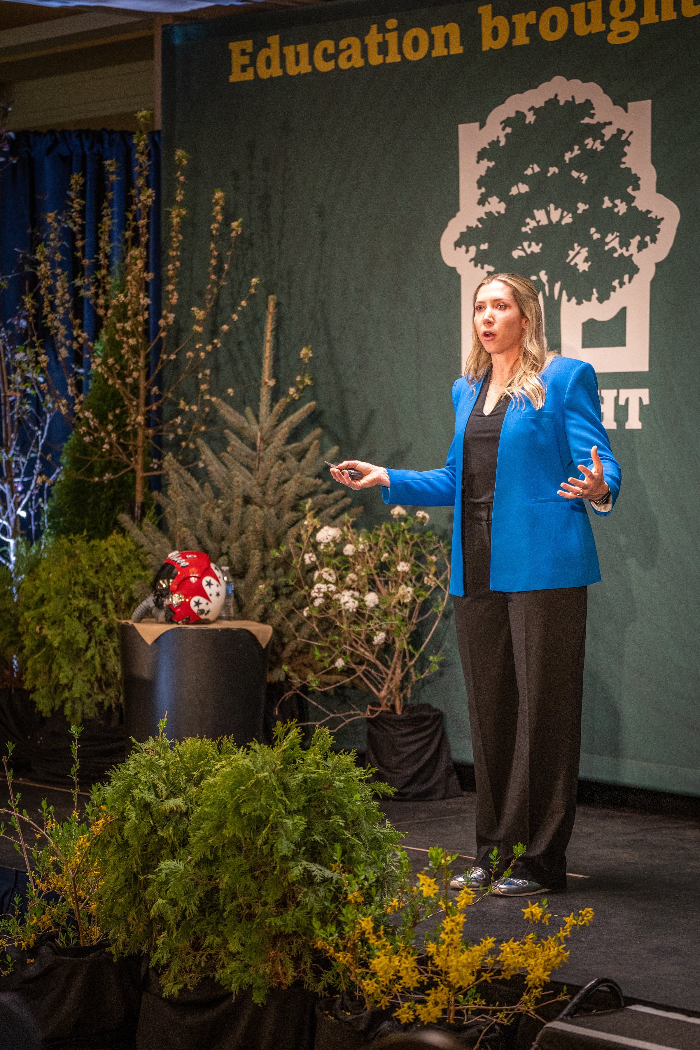 A woman in a blue blazer and black pants talks on a stage decorated with various potted plants and yellow flowers. She holds a remote in her right hand. Behind her, a large banner reads "Education brought" with a logo of a tree. A red helmet with whi