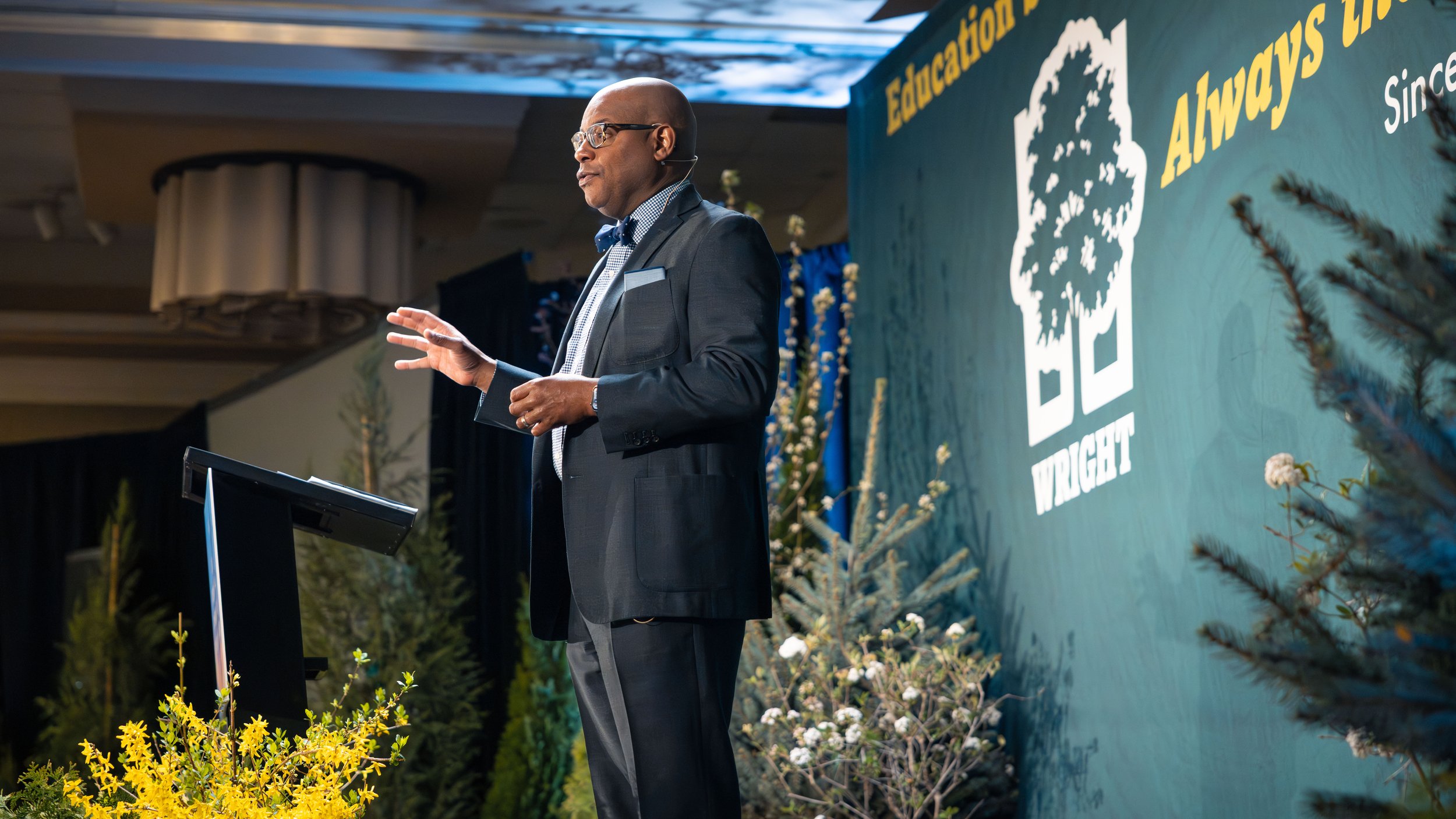 A man in glasses and a suit is giving a presentation on stage, with a backdrop that reads 'Education' and 'Always' along with a logo, surrounded by plants and flowers.