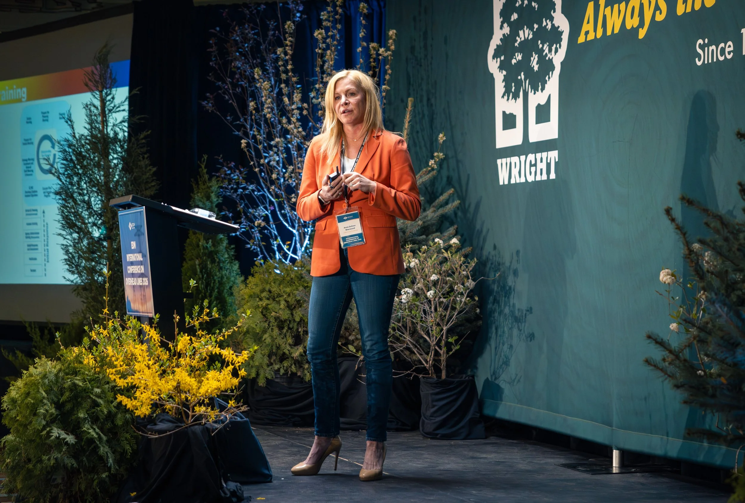 A woman with long blonde hair wearing a rust-colored blazer, white blouse, and dark blue jeans, giving a presentation on stage with a lectern, surrounded by trees and plants, at a conference with a large screen and a banner that reads 'Always the Bes
