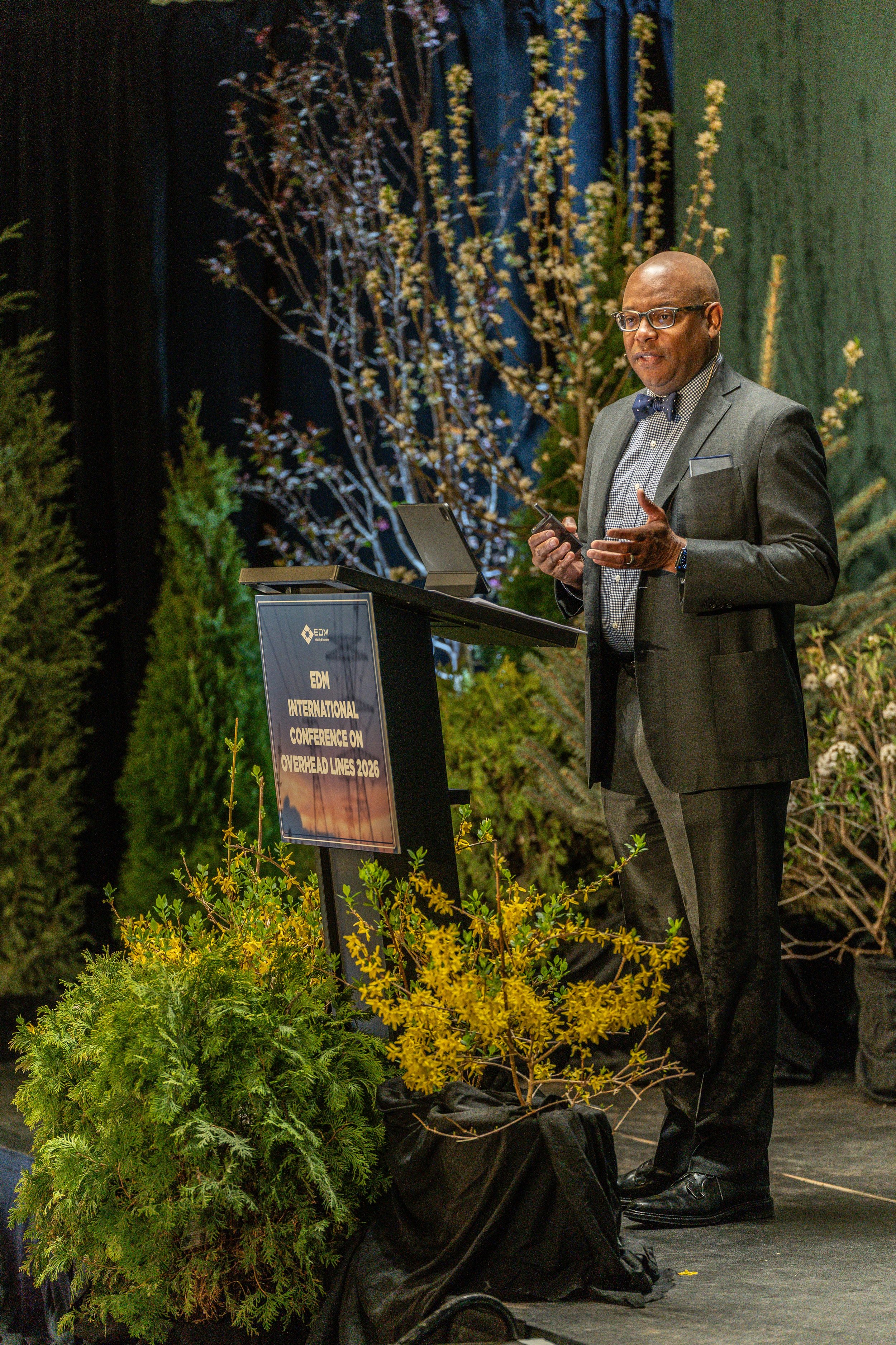 A man in a gray suit, checkered shirt, and bow tie standing in front of a floral backdrop, giving a presentation at the EDM International Conference on Overhead Lines 2026. There is a lectern with a sign and plants in the foreground.