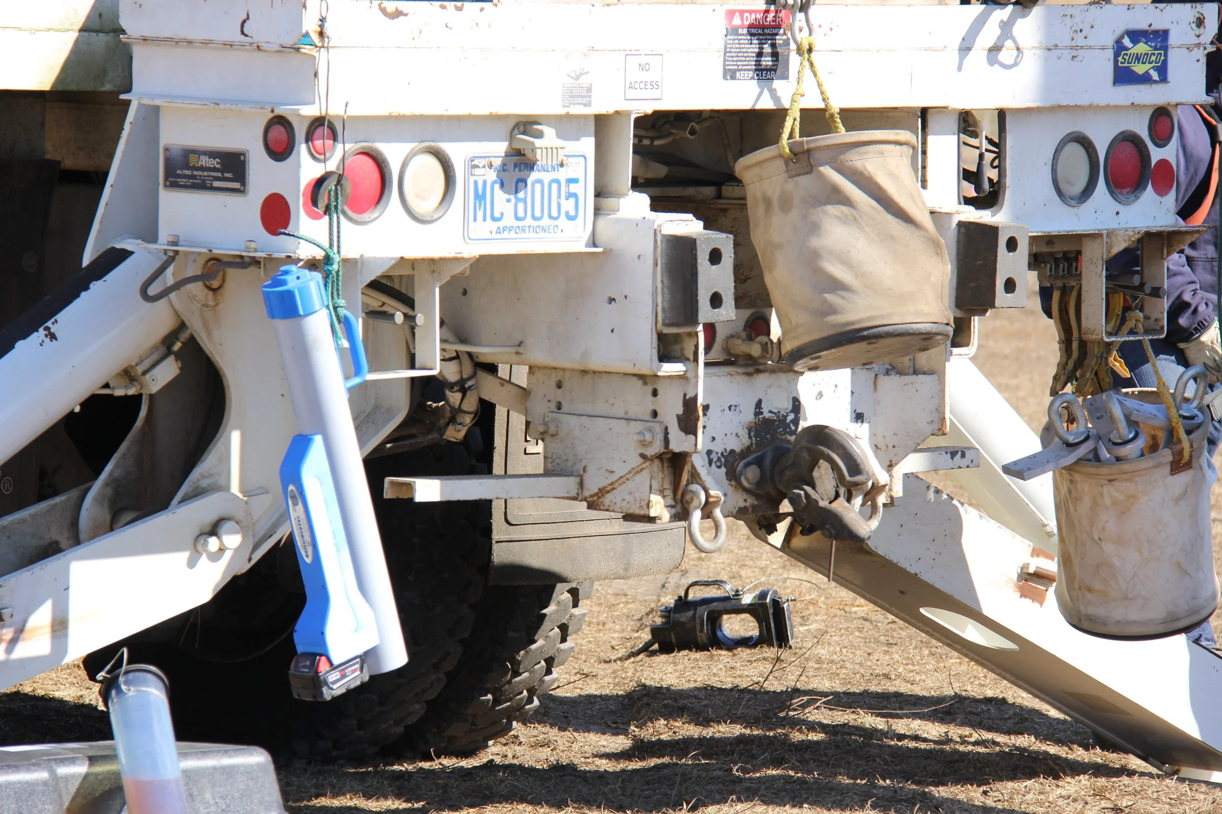 Close-up of the back of a utility vehicle with tools, bags, and equipment hanging and resting on it, including a bucket and a wrench, on a dirt ground.
