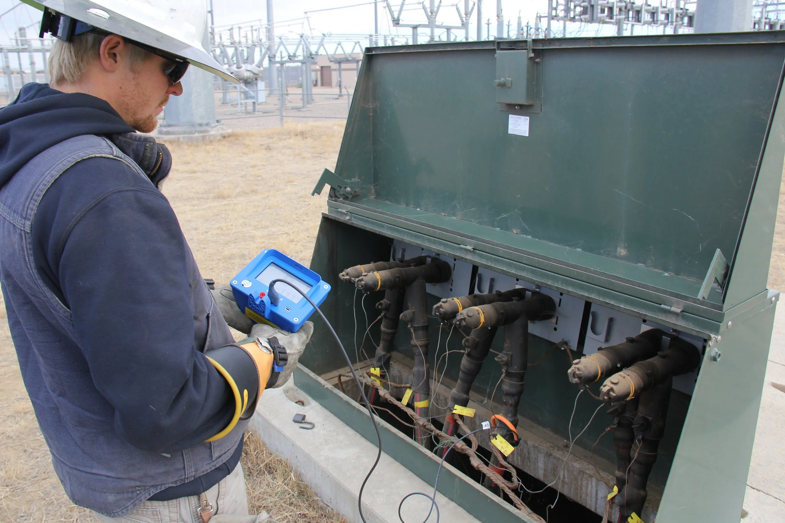 A technician wearing a white helmet and gloves uses a handheld testing device on electrical equipment in an outdoor power station.