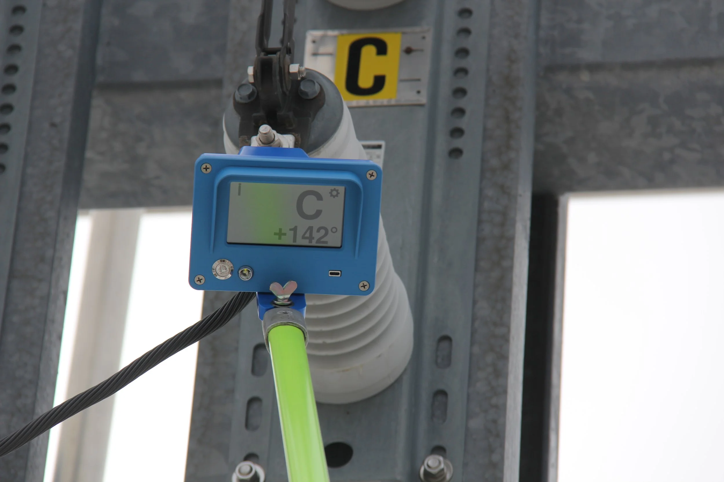 Close-up of a digital device attached to an electrical pole, displaying a reading of +142 degrees. The device is connected to a green cable, with a background of metal electrical infrastructure.