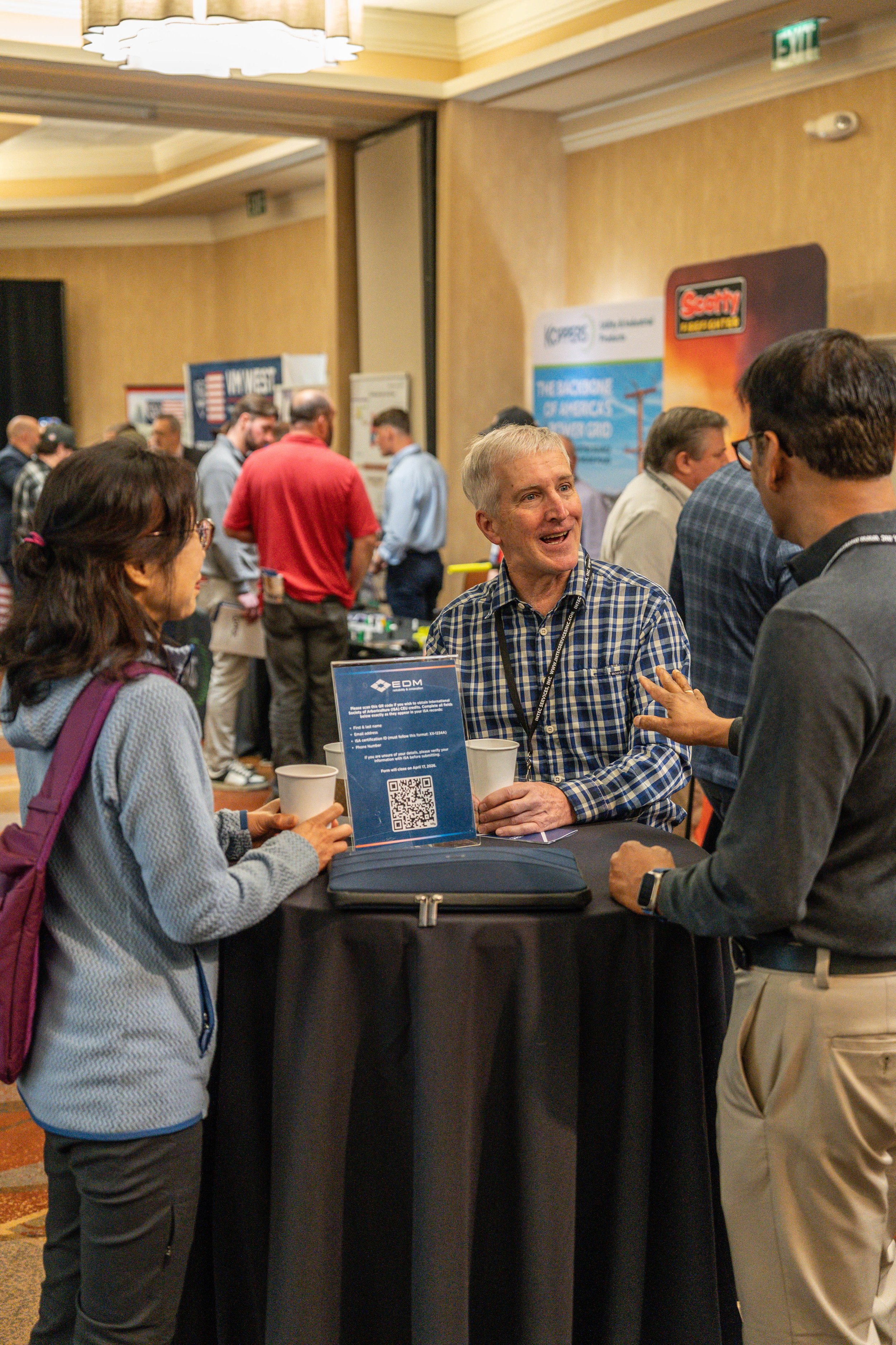 A group of people at a convention or conference engaging in conversations. There are banners and posters in the background, and a person is speaking to another at a round table with informational materials.