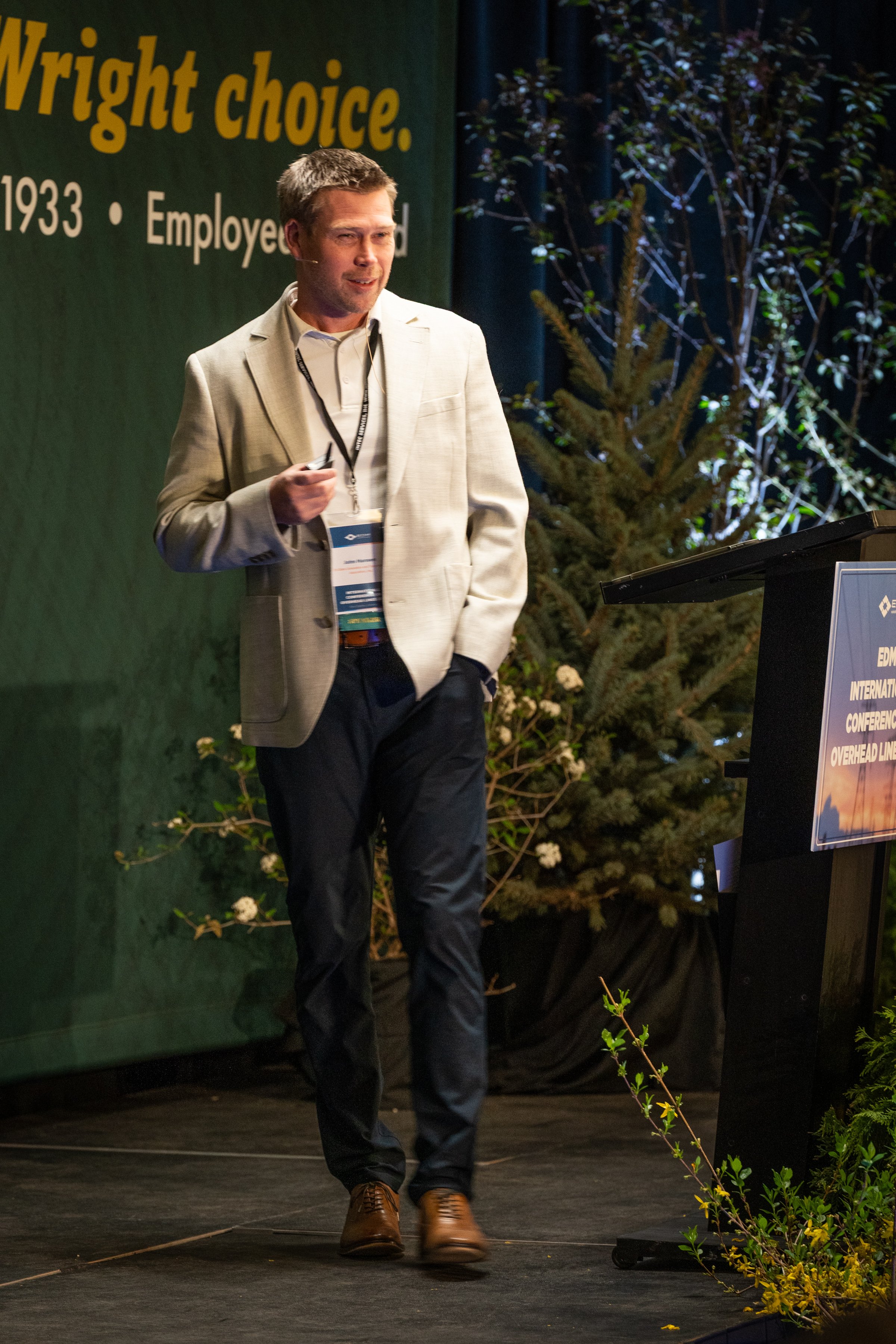 A man in a light-colored suit jacket and dark pants giving a presentation on stage, standing near a lectern with a screen and a large green backdrop with yellow text, and some foliage and plants in the background.