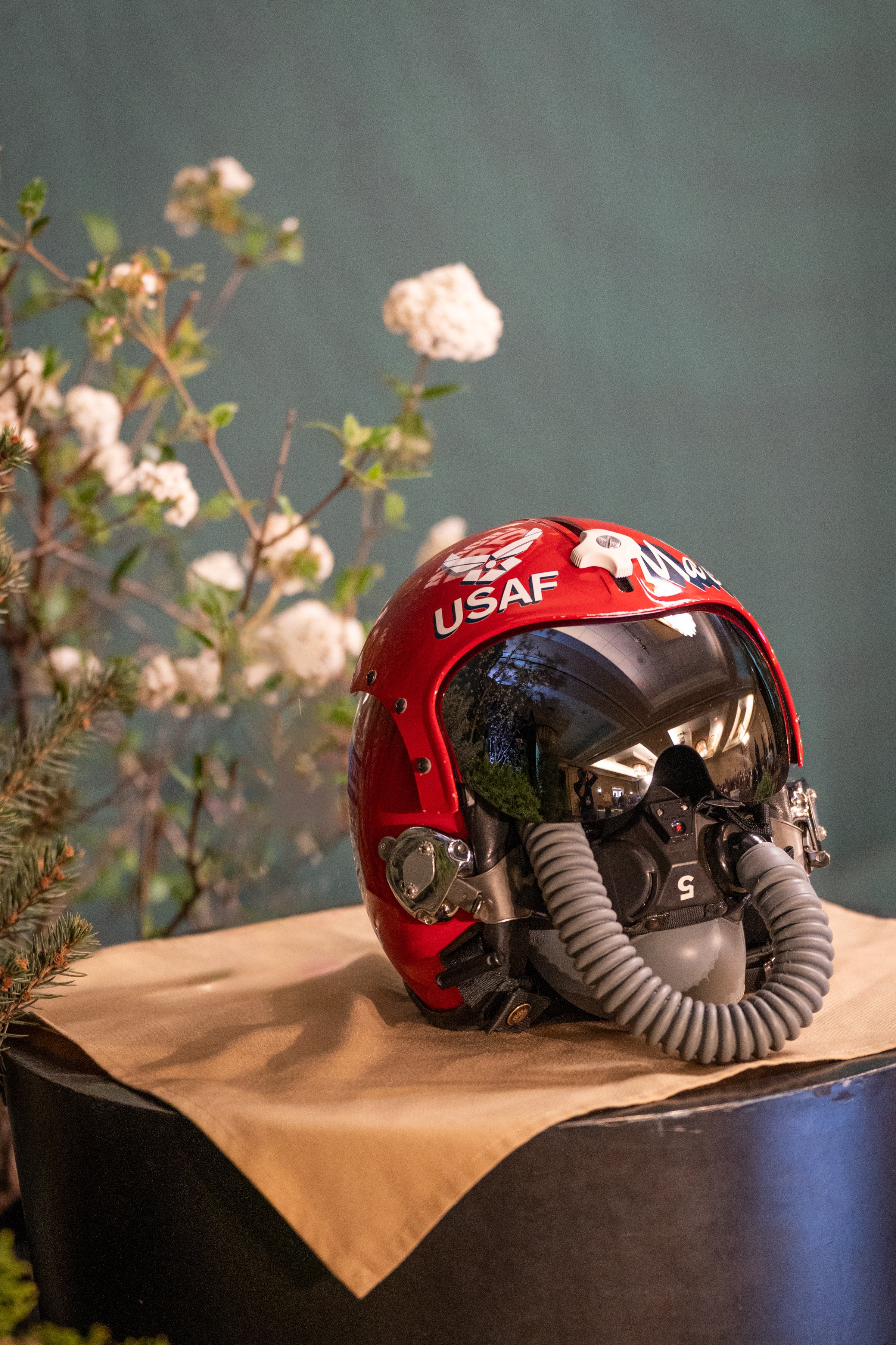 A red aviation helmet with a visor and the USAF logo, placed on a surface with a piece of brown paper, with some white flowers and green leaves in the background.