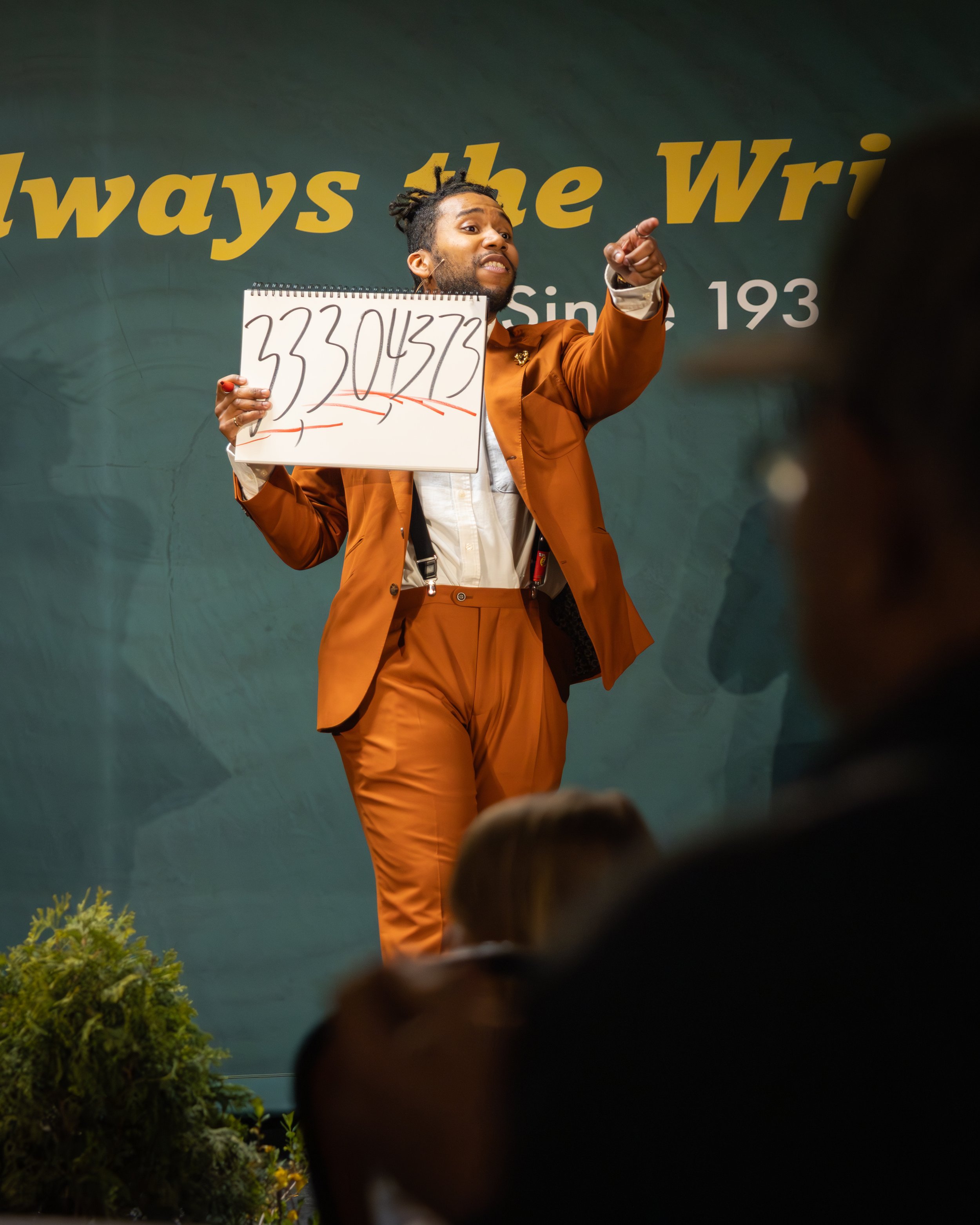 A man in a brown suit pointing and speaking during a presentation or event, holding a whiteboard with the number 33304373 written on it, with greenery and a dark background behind him.