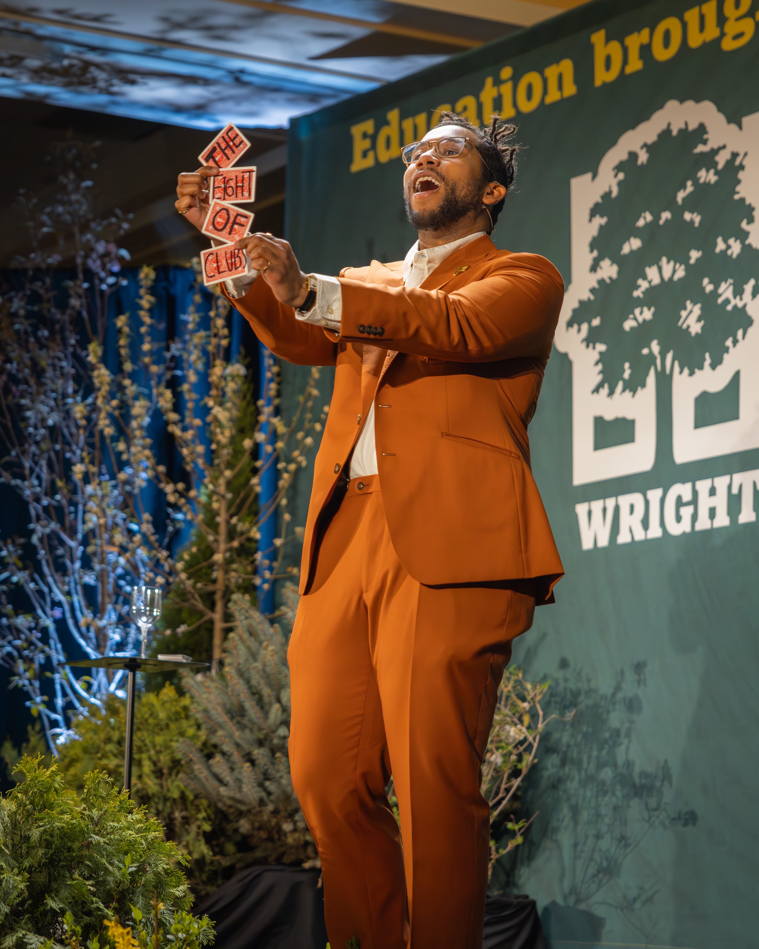 A man wearing glasses dressed in an orange suit speaking on stage at an outdoor event, holding cut-out signs that spell 'THE FIGHT OF CLUB,' with a backdrop that reads 'Education' and a scene of nature with trees and plants.