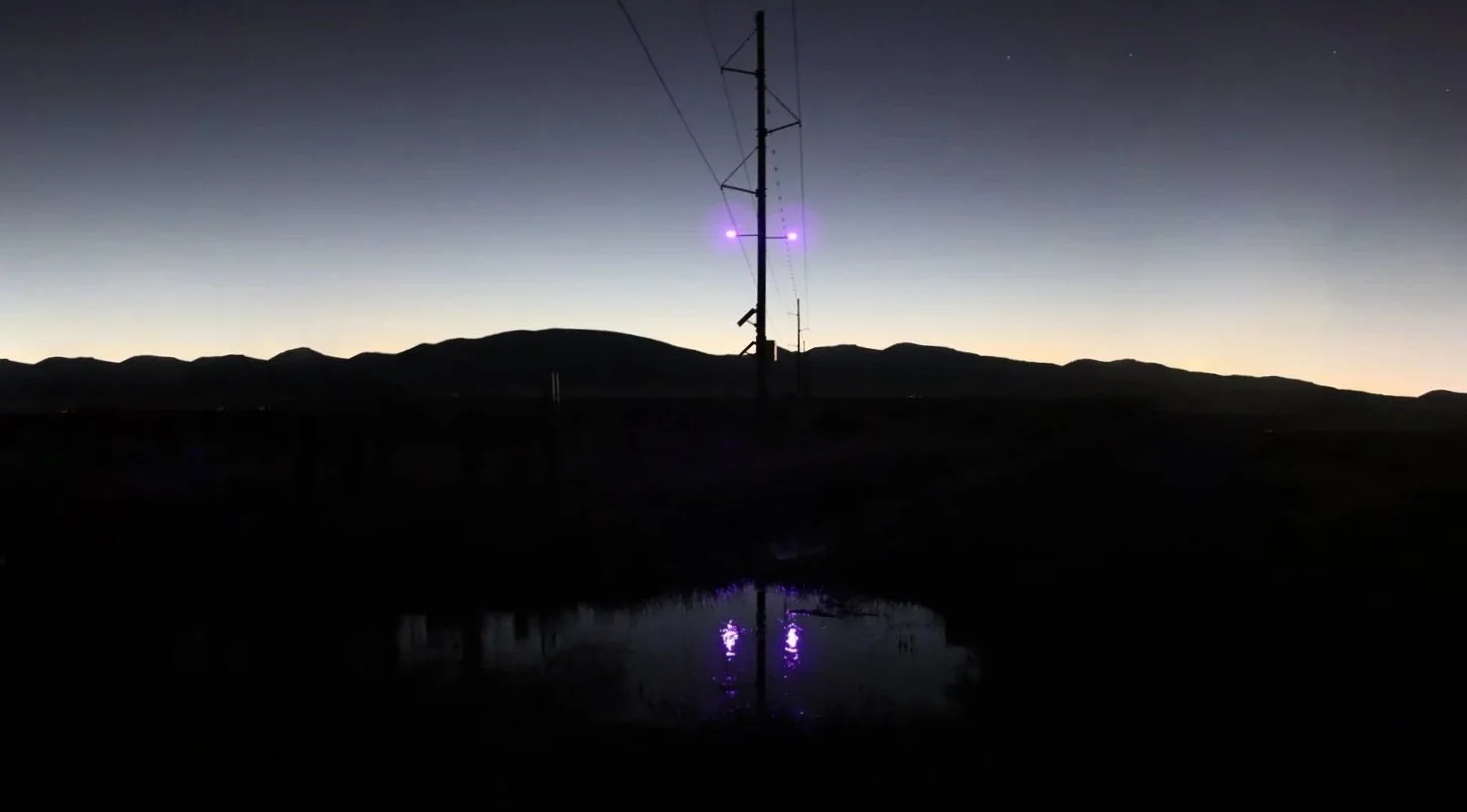 A silhouette of a power line pole and wires against a twilight sky, with mountains in the background and a small body of water reflecting purple lights from the pole.
