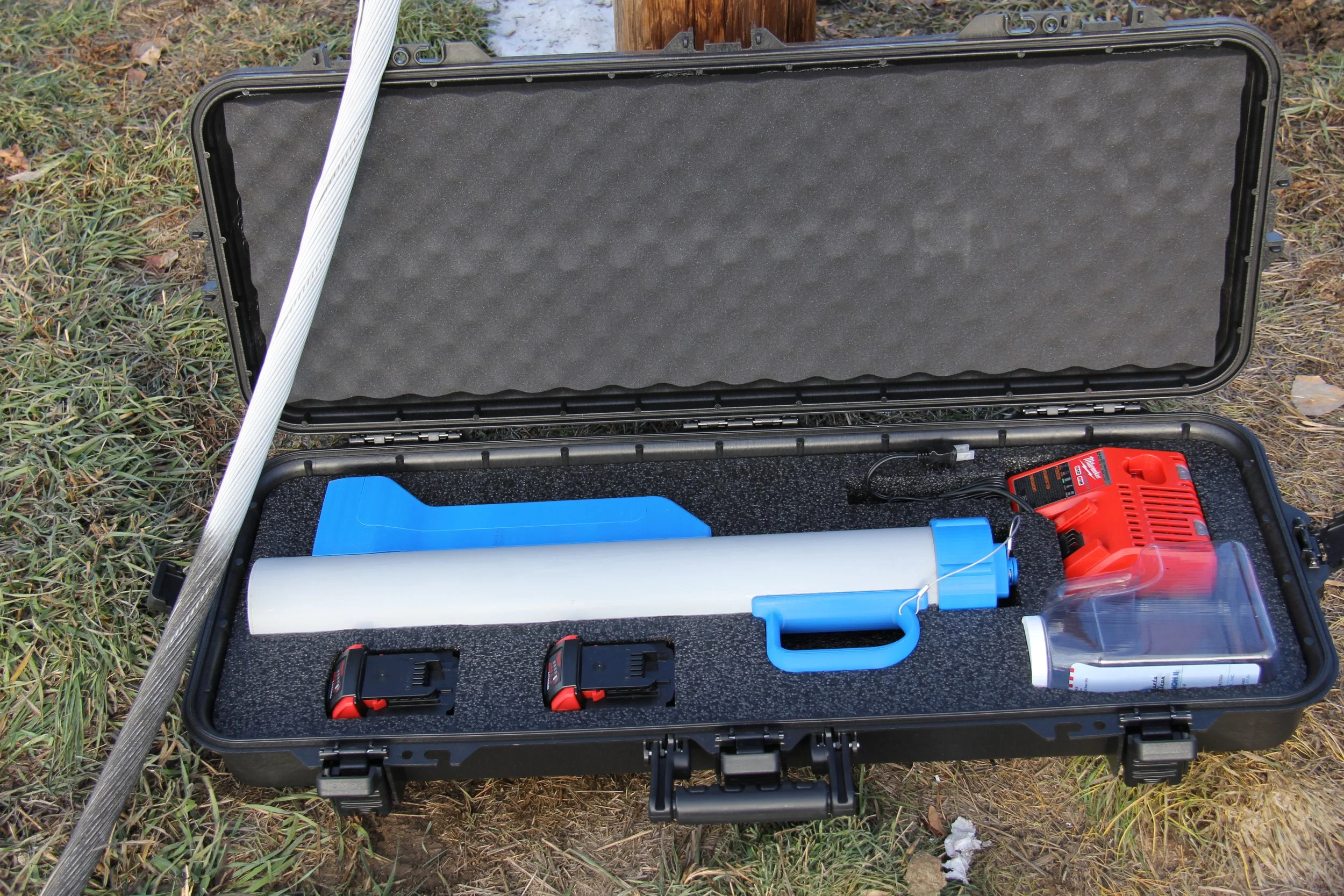 A black carrying case with foam interior, open outdoors on grass, containing a long white tube, a blue handle, two battery packs, a red power tool, and a plastic water bottle with some paper beside it.