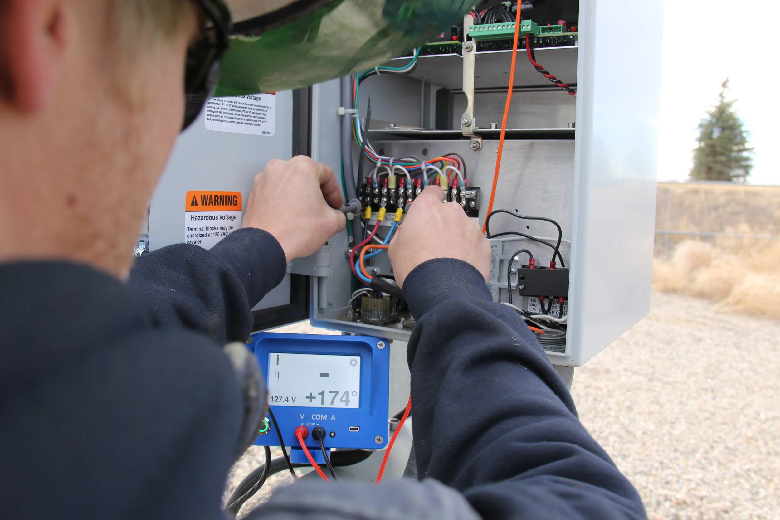 A technician working on an electrical control panel outdoors, with a multimeter measuring voltage.