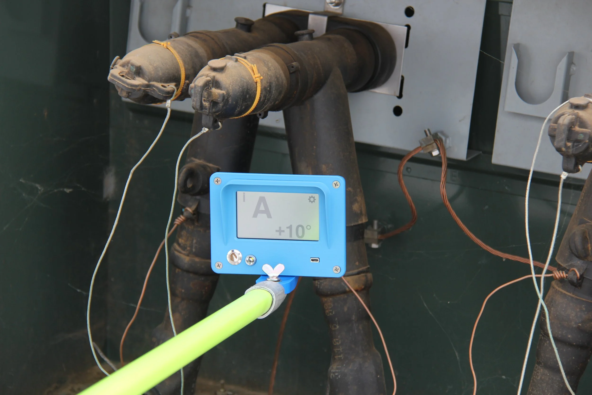 A pair of large, cylindrical, black and rusted industrial components mounted on a metal stand, with wires attached. A blue digital device displaying '+10°' and 'A' is held by a person wearing a high-visibility glove in front of the components.