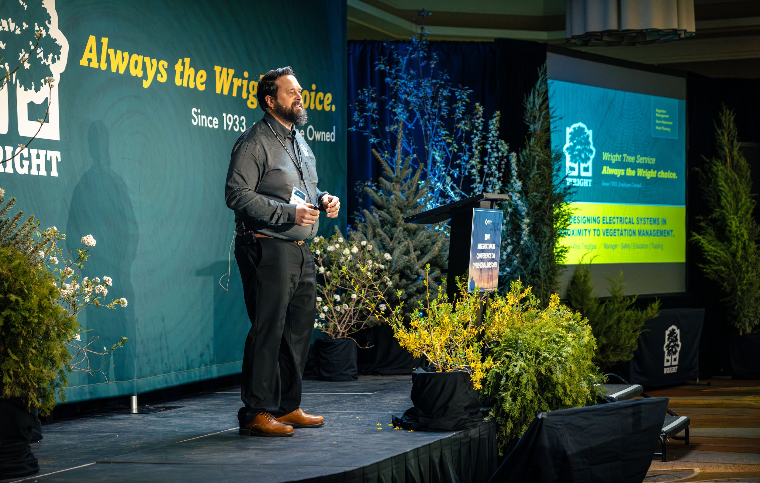 Man with beard and dark hair wearing gray shirt and brown shoes standing on stage with a microphone, trees, and a large screen displaying green and blue graphics behind him at an indoor event.