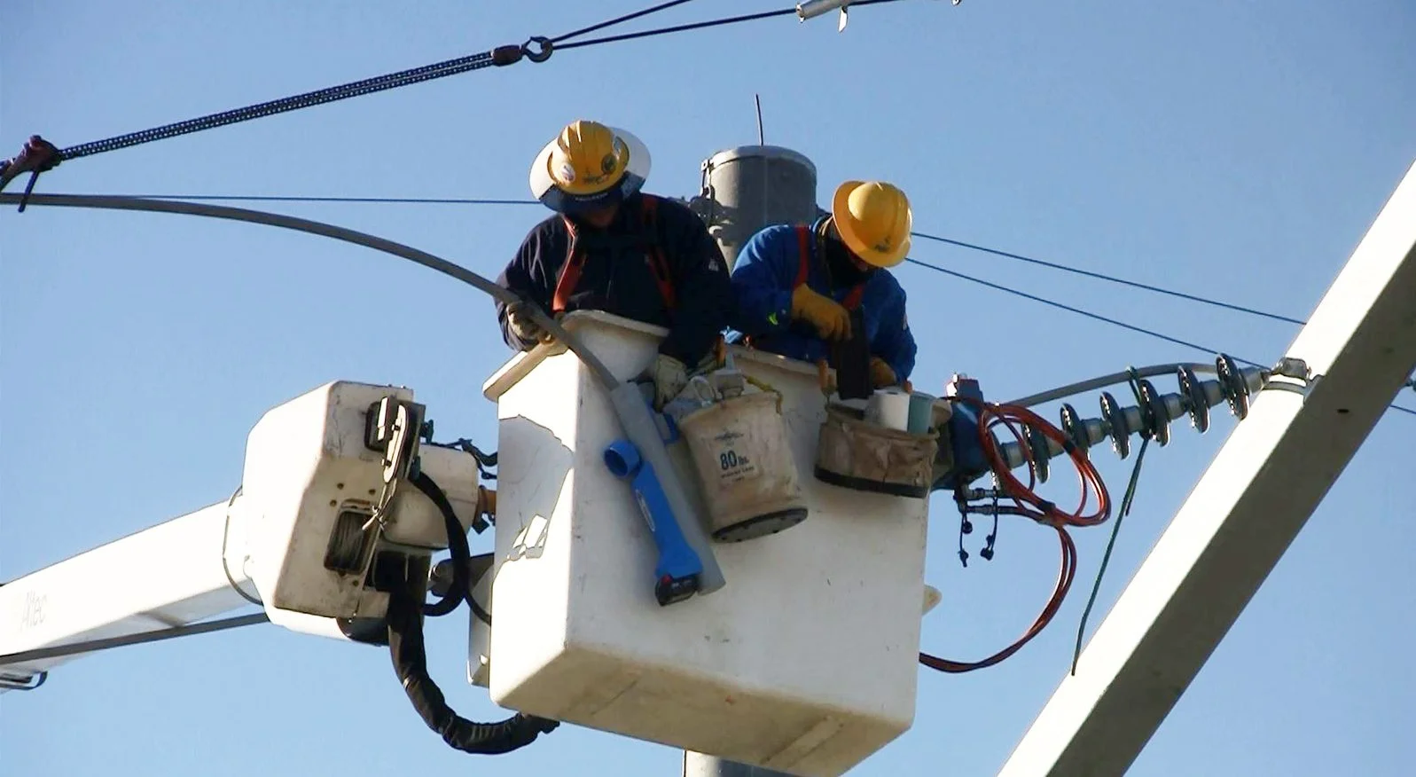Two utility workers wearing yellow safety helmets working inside a white bucket lift on electrical power lines against a clear blue sky.