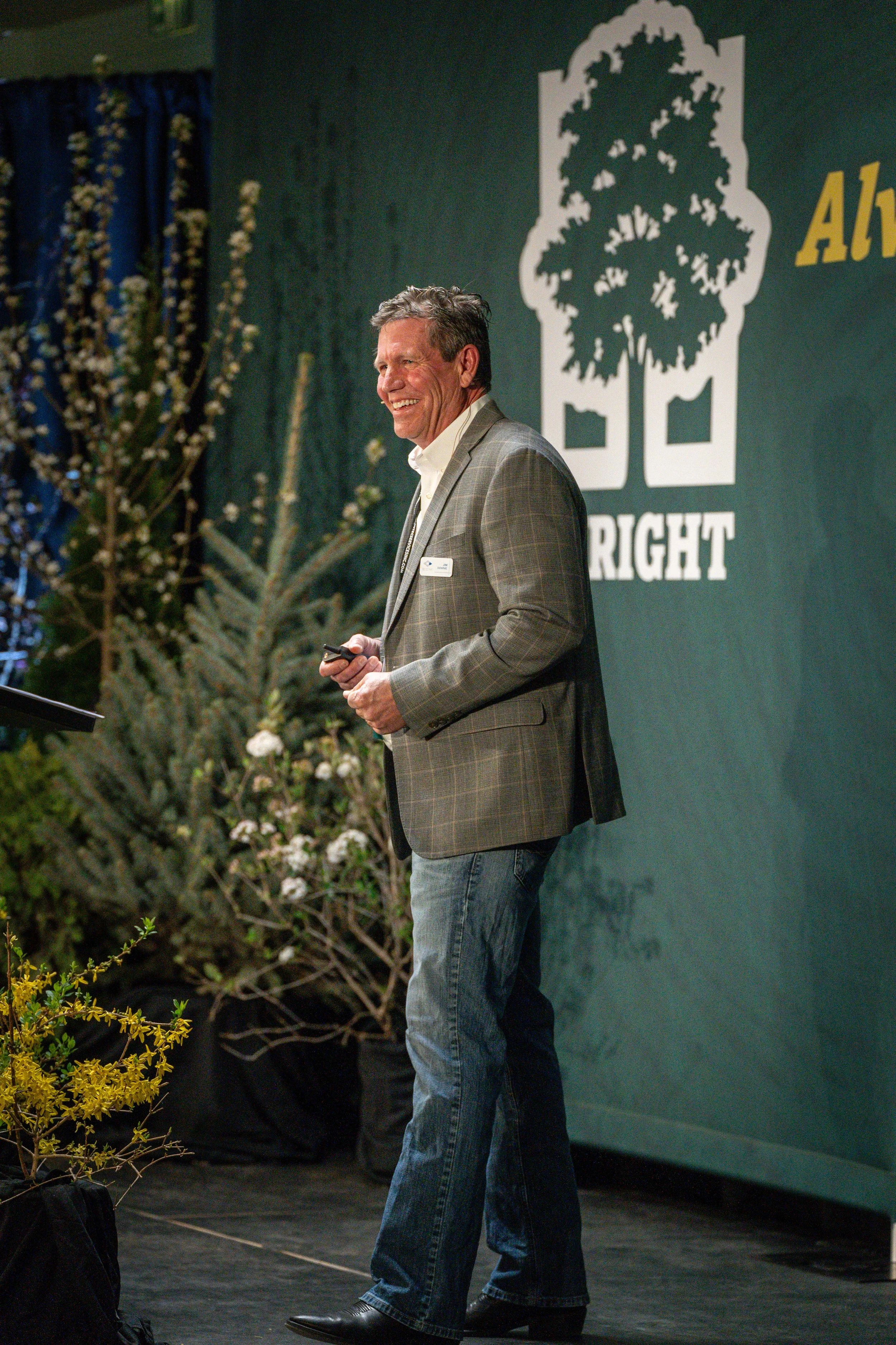 A man with gray hair wearing a gray plaid blazer, white shirt, and jeans standing on a stage, smiling, holding a small device, with a large green backdrop featuring a white tree and the words 'All the Right' and a partial yellow logo on the right. Th
