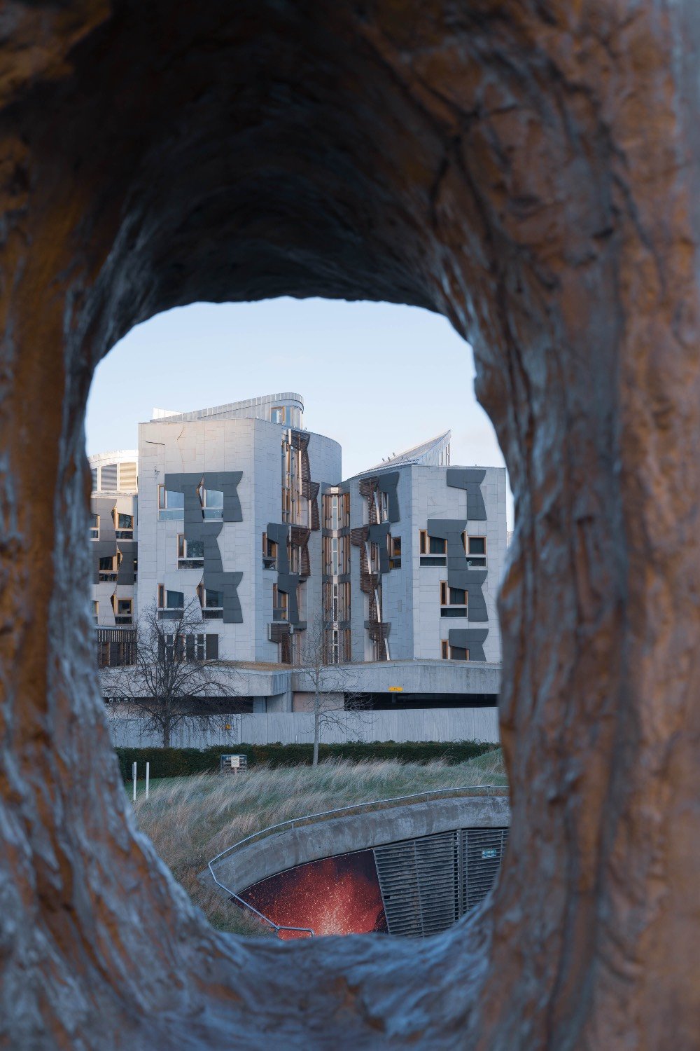 The modern geometric windows of the Scottish Parliament building framed through a circular opening in a weathered bronze sculpture.