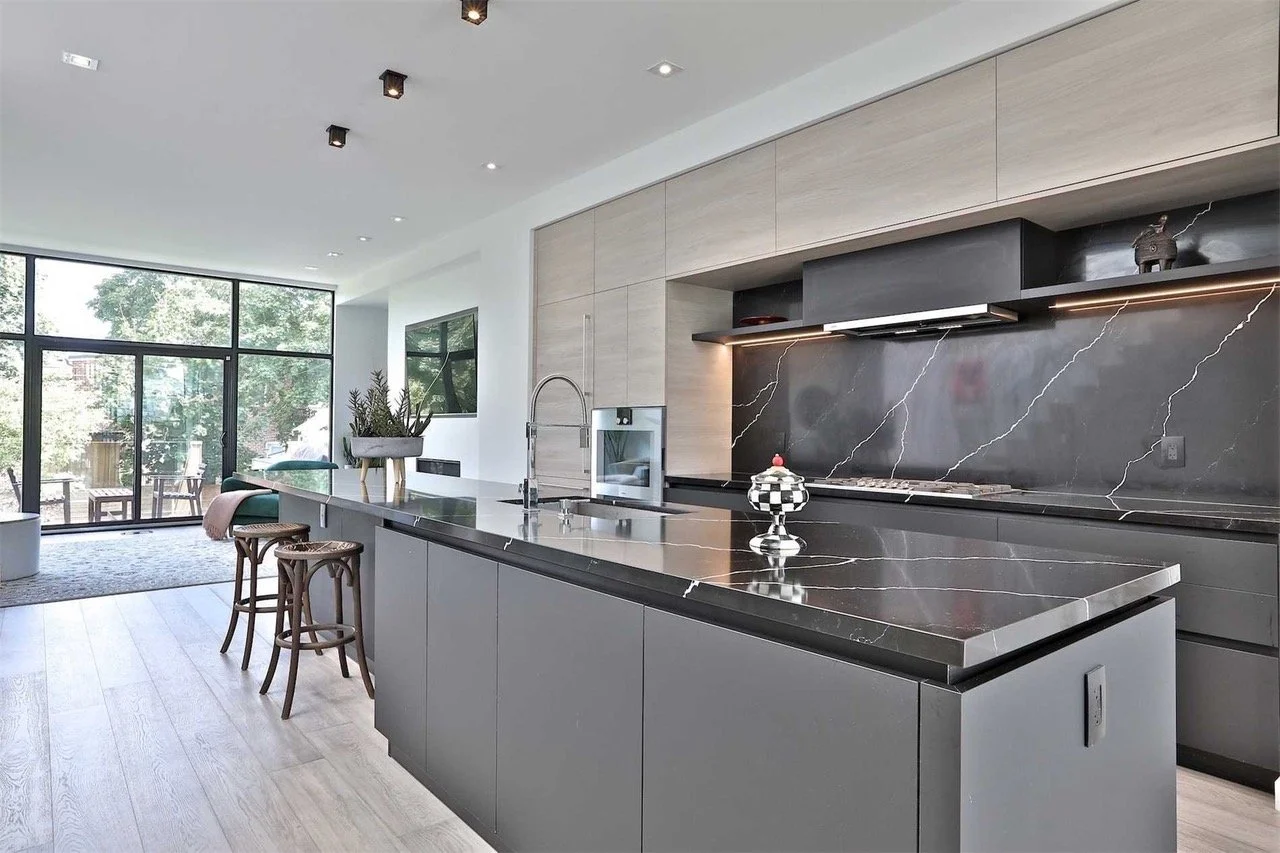 Modern kitchen with black marble countertops, gray cabinets, and a black marble backsplash with white veining. Large window views of greenery outside.