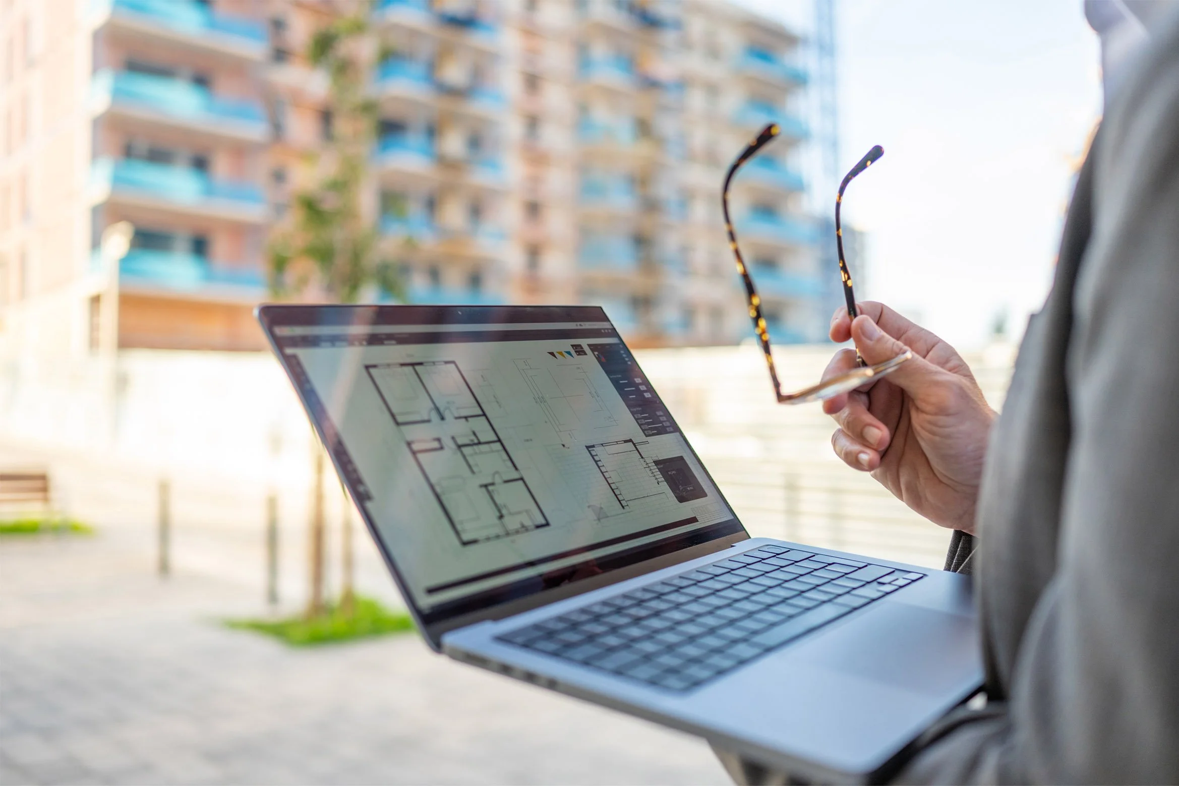 Person holding glasses and working on architectural blueprints on a laptop outside with high-rise buildings in background.