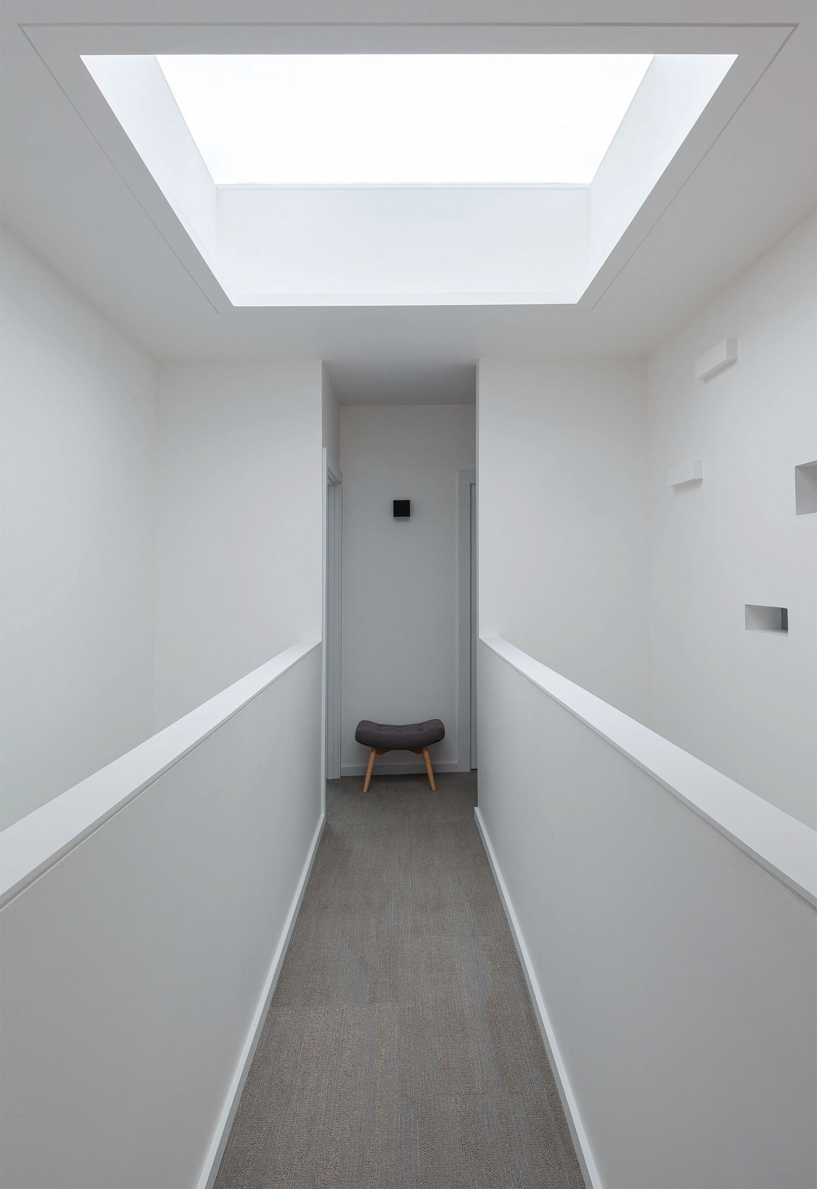 A narrow, white hallway with a skylight overhead, a small cushioned stool at the end, and minimal decor.