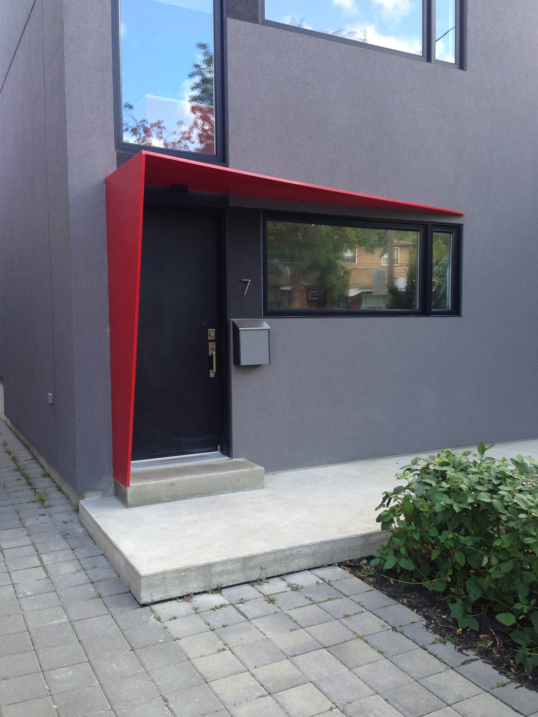 Whale House entrance in East York, Toronto, built by Maxamin Homes, featuring black door, red awning, black-framed windows, and green bushes.