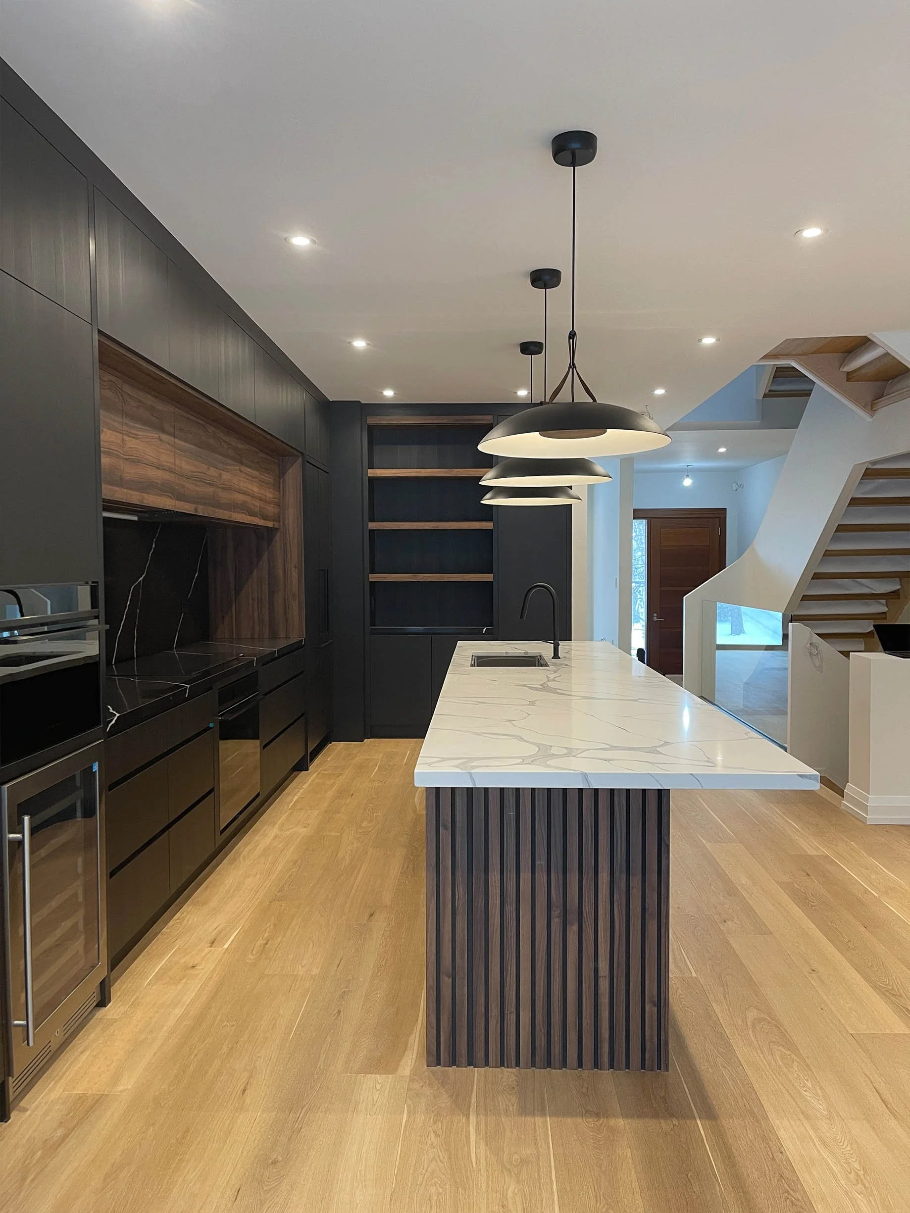 Modern kitchen with black cabinetry, a white marble island, wooden floors, and a staircase in the background.