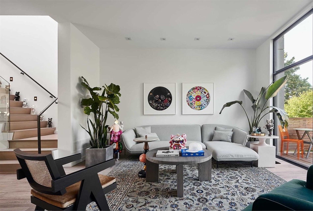 Modern living room with white walls, a large gray sectional sofa, and a round coffee table with toys on it. There are two colorful circular art pieces on the wall, large plants near the sofa, and large windows with a view of trees outside. A staircas