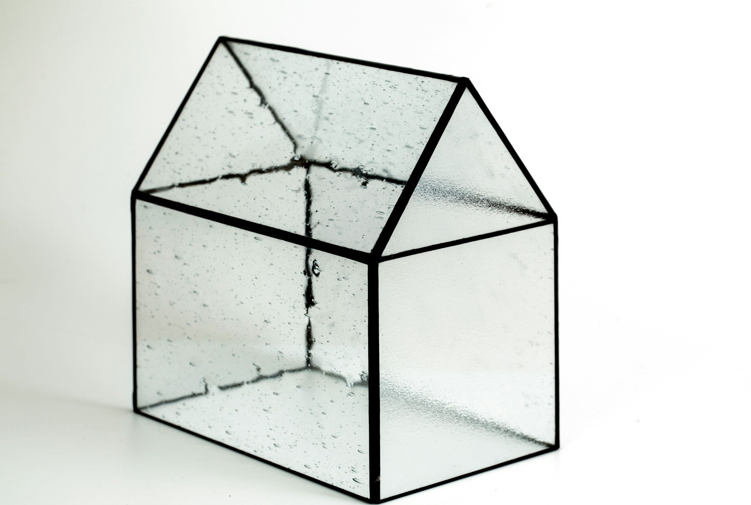 A 3D geometric sculpture resembling a house, made of transparent glass with black outlines, featuring water droplets inside.