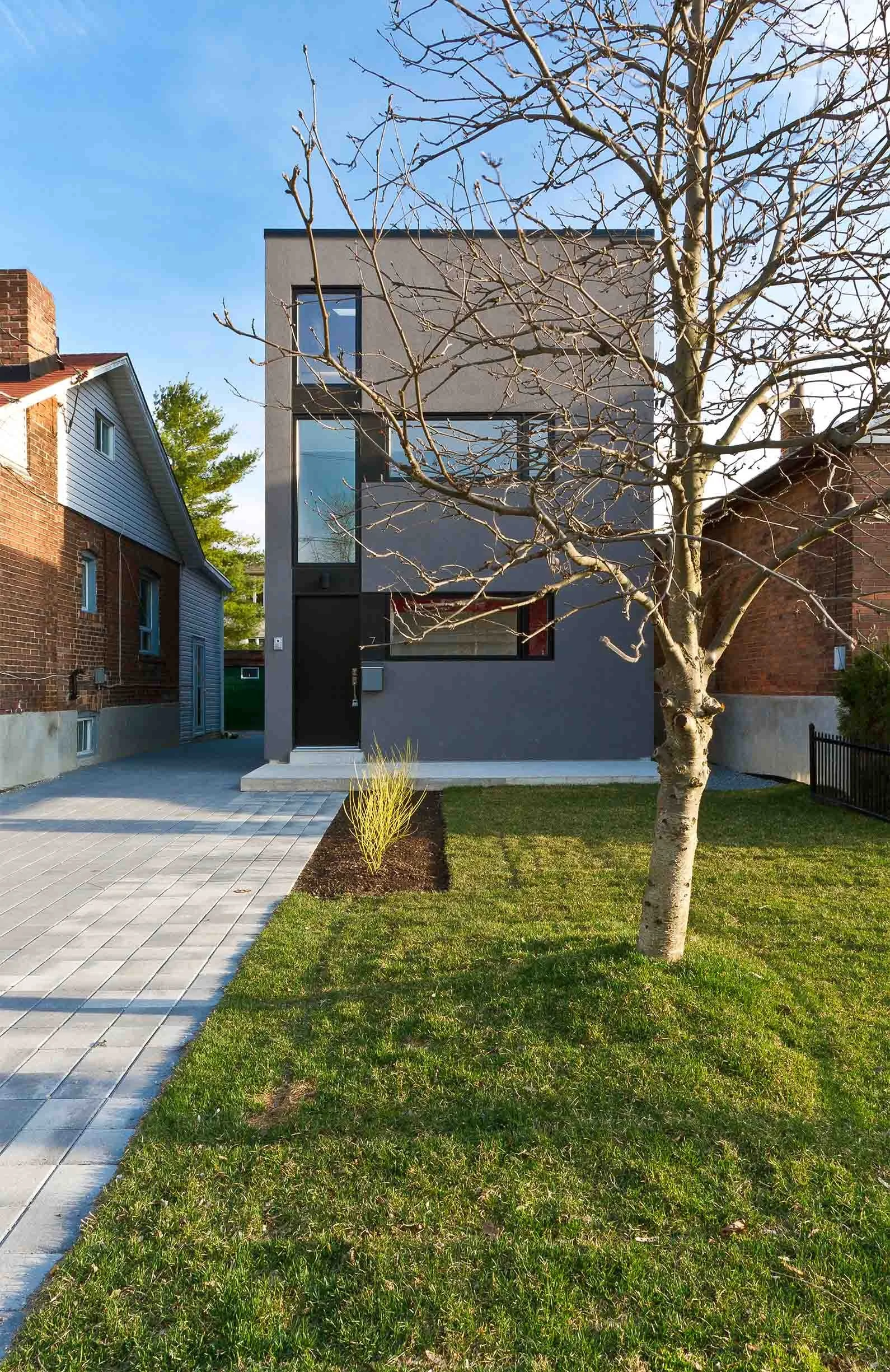 Whale House exterior in East York, Toronto, built by Maxamin Homes, gray three-story home with large windows, small lawn, tree, and paved walkway.