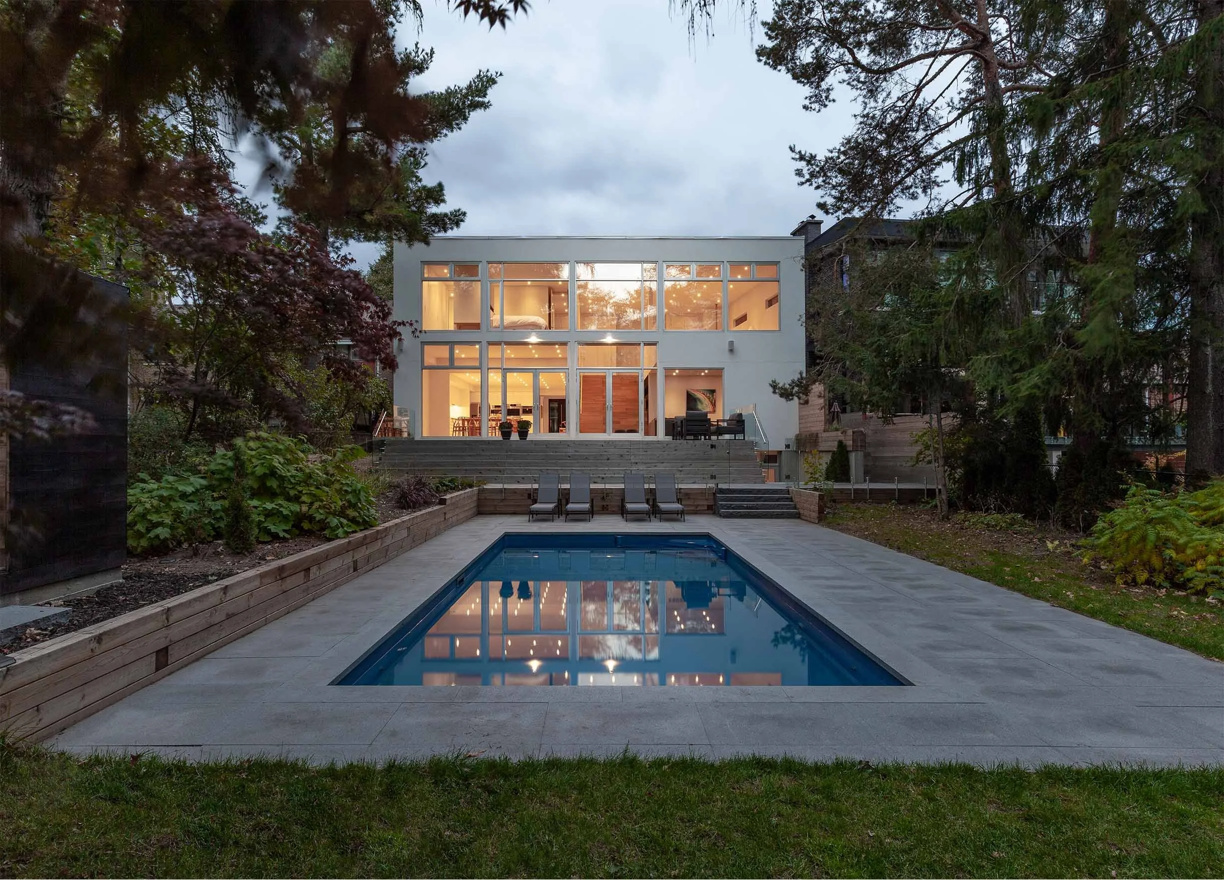 Fun House, a modern luxury custom home in East York, Toronto, built by Maxamin Homes, featuring large glass windows, an outdoor swimming pool, and a landscaped yard illuminated in the evening.