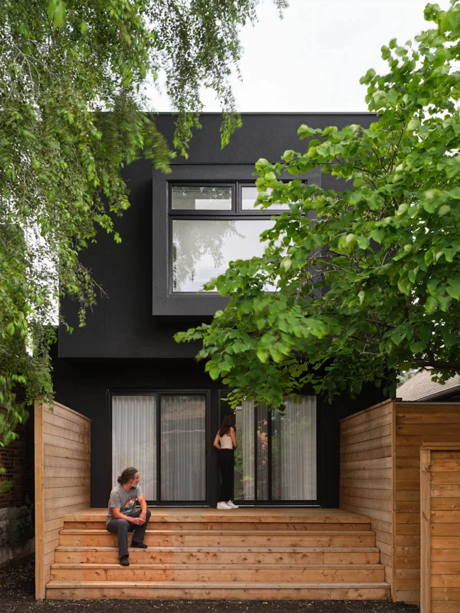 Periscope House backyard in East York, Toronto, built by Maxamin Homes, showing black two-story exterior, wood deck, and people enjoying outdoor space.