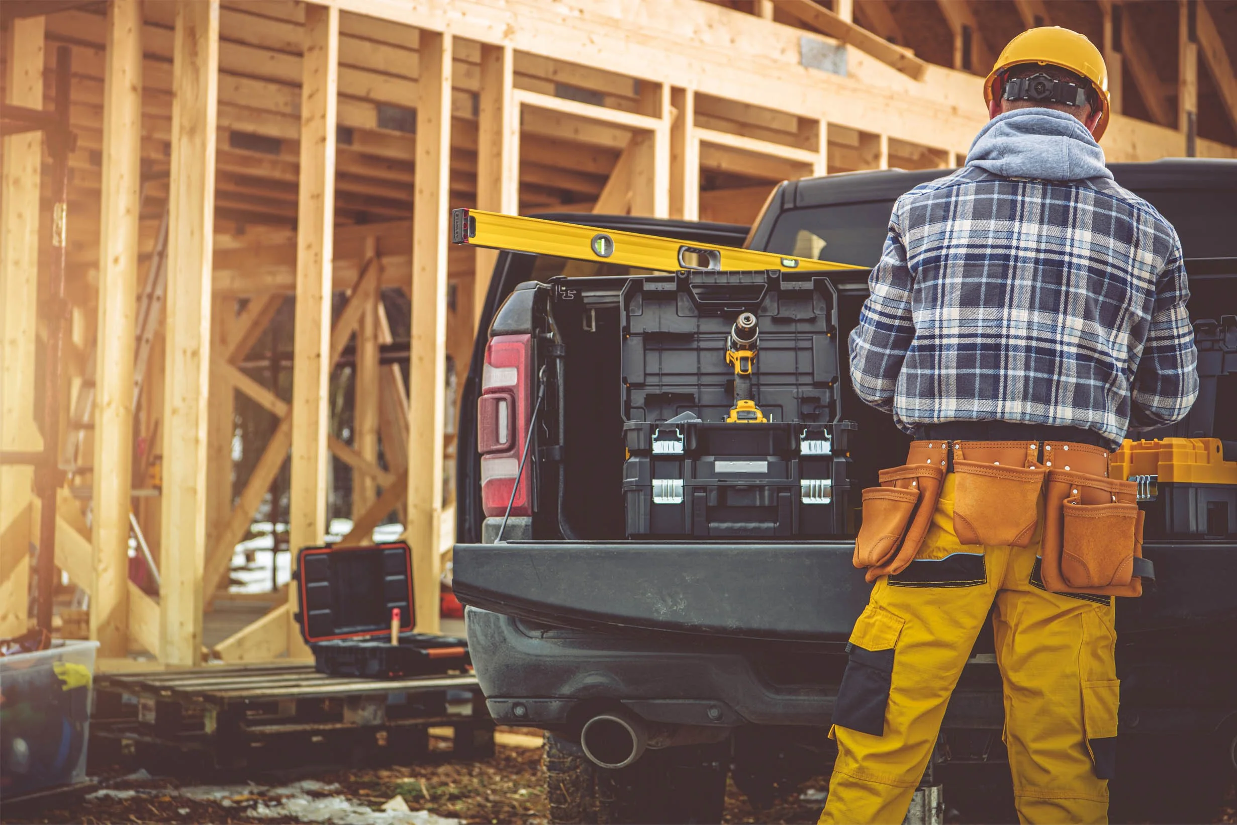 Construction worker wearing yellow pants, a plaid jacket, and a yellow safety helmet standing at the back of a pickup truck filled with tools at a building site with wooden framing.