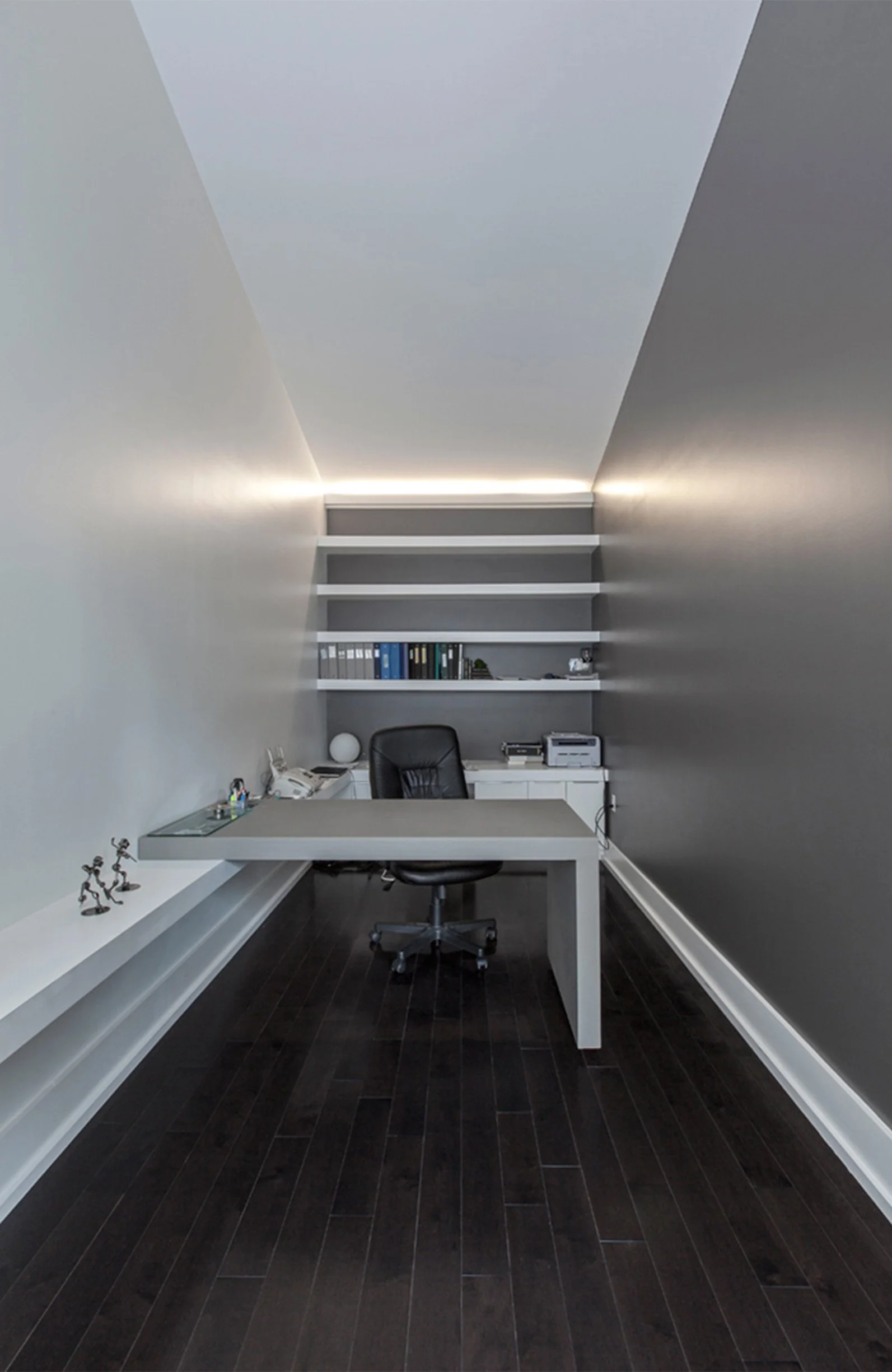 Minimalist modern office with a white desk, black swivel chair, dark wood floor, white shelves with binders and decorative items, and gray walls with ambient lighting.