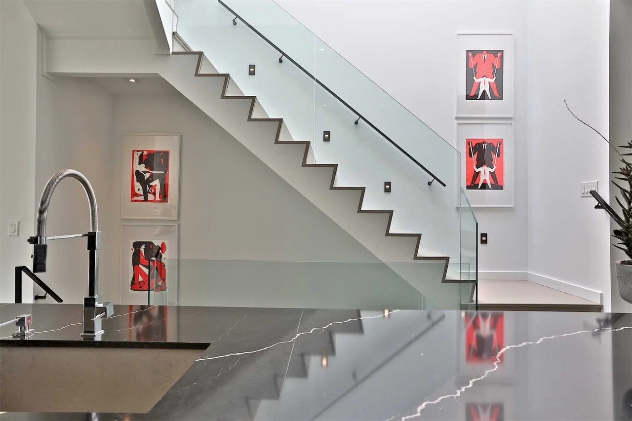 Modern interior space with a staircase, glass railing, and three framed abstract art pieces on the wall featuring red, black, and white colors. Part of a black marble kitchen counter with a sink is visible in the foreground.
