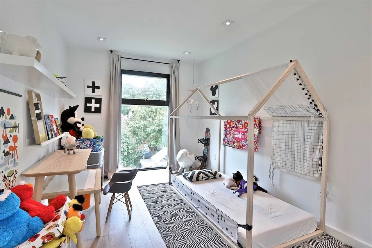 A child's bedroom with a house-shaped bed frame, plush toys, wall art, a window with curtains, and a desk with a chair, in a modern and playful decor style.