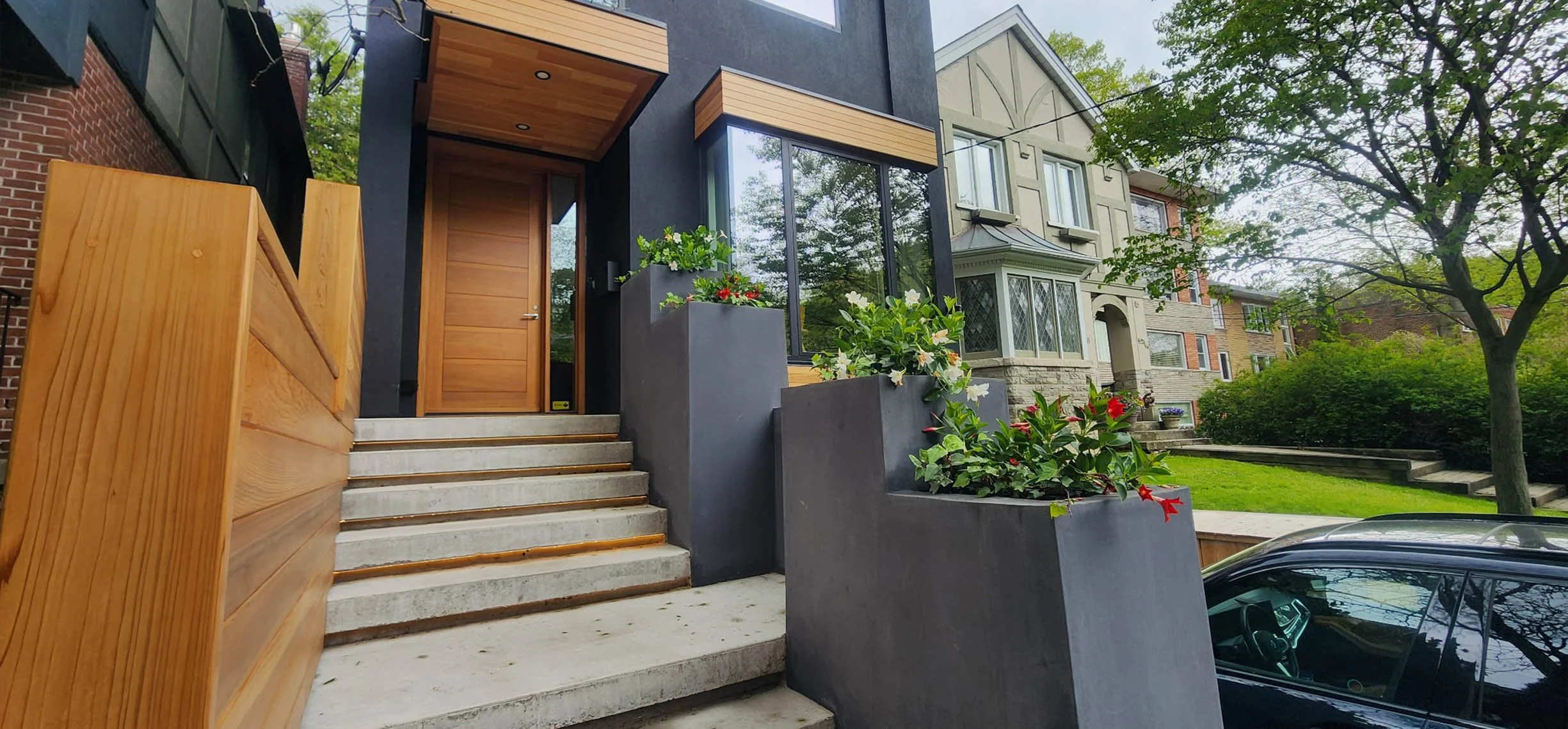 Lantern House entrance in The Beaches, Toronto, built by Maxamin Homes, featuring concrete steps, wooden door, and potted plants by flowering bushes.