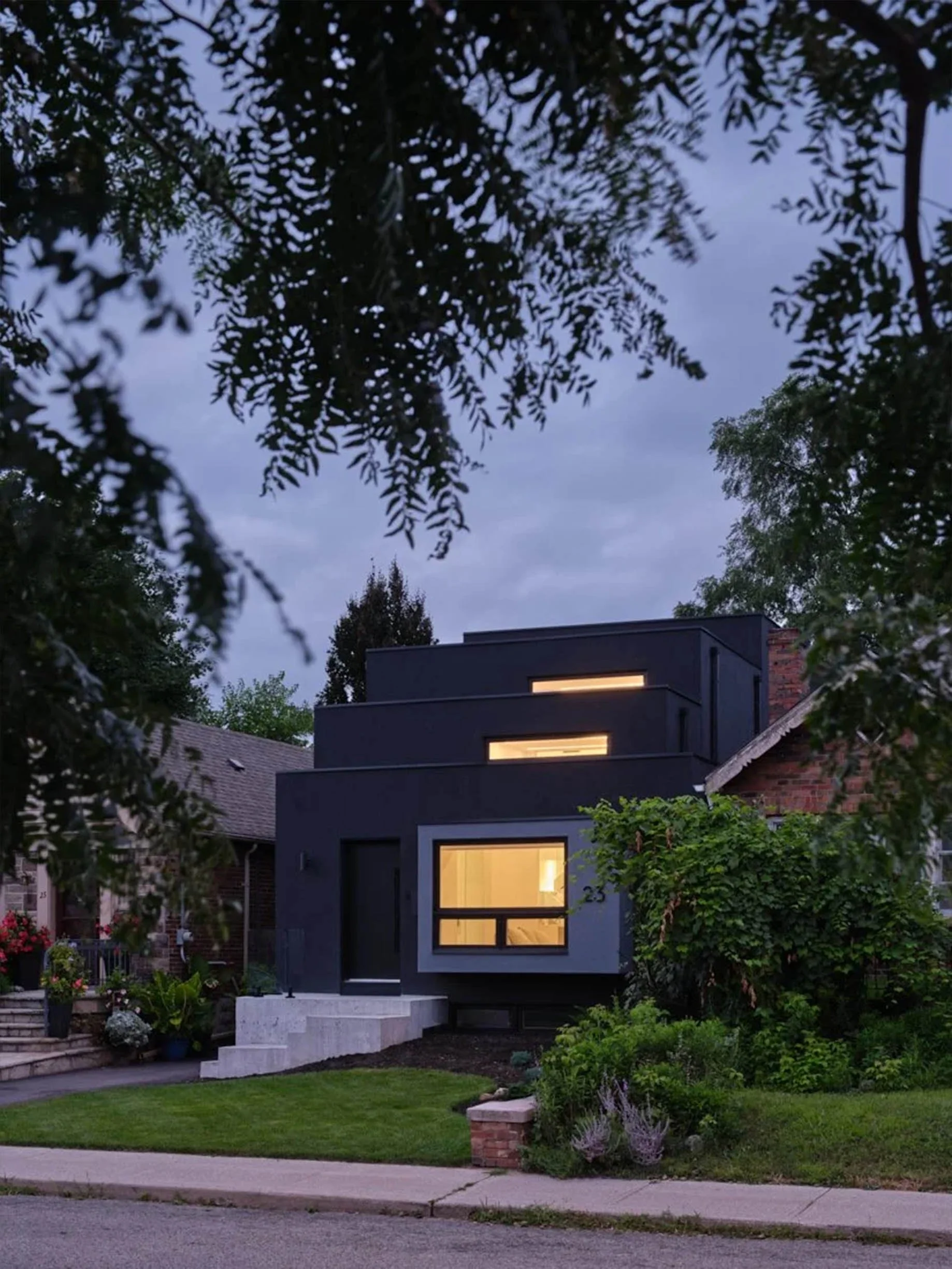 Periscope House exterior in East York, Toronto, built by Maxamin Homes, modern black home illuminated at dusk among greenery and nearby houses.