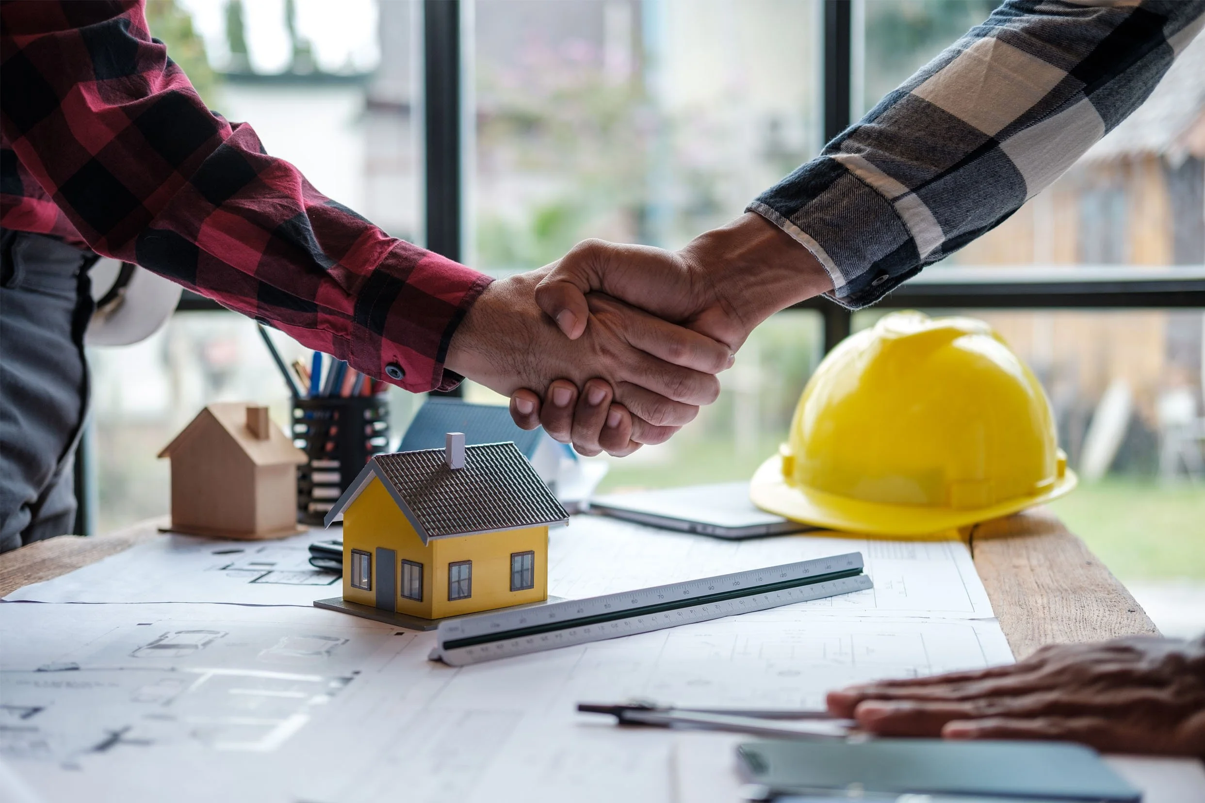 Two people shaking hands over a table with architectural plans, miniature houses, a yellow construction helmet, and a ruler, in front of a large window.